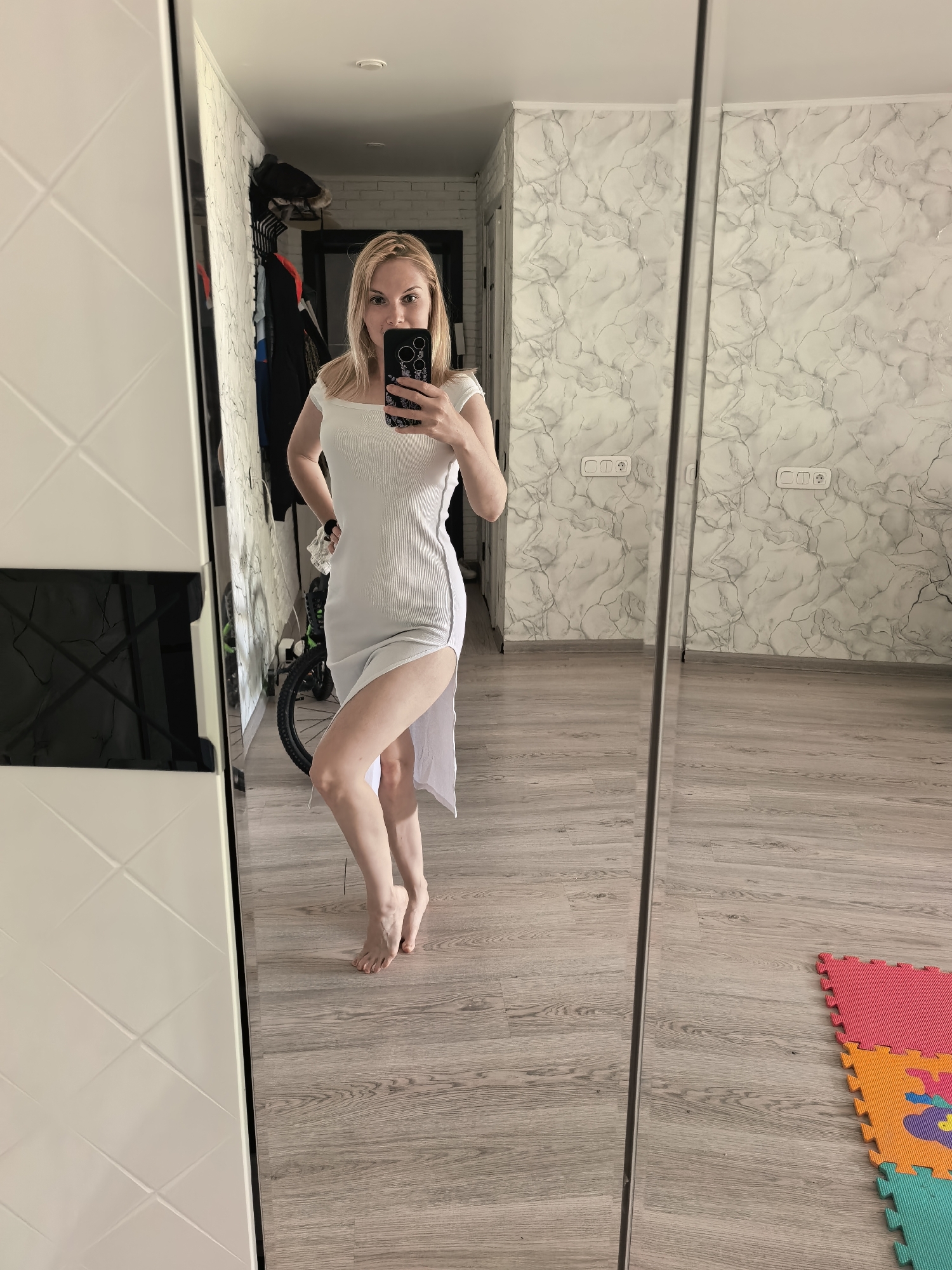 A fair-skinned woman with blonde hair is taking a mirror selfie in what appears to be her home. She is wearing a white, off-the-shoulder dress with a high slit. She holds a smartphone in her right hand, partially obscuring her face. The background includes a closet with white and black panels, a bicycle, and a doorway leading to another room with white brick walls. The wall visible to the right has a marble-like pattern. The floor is light-colored wood. In the bottom right corner, a colorful children's play mat is visible. Given the presence of domestic objects and the typical interior design, the image likely originates from a private residence in Sterlitamak, Russia.