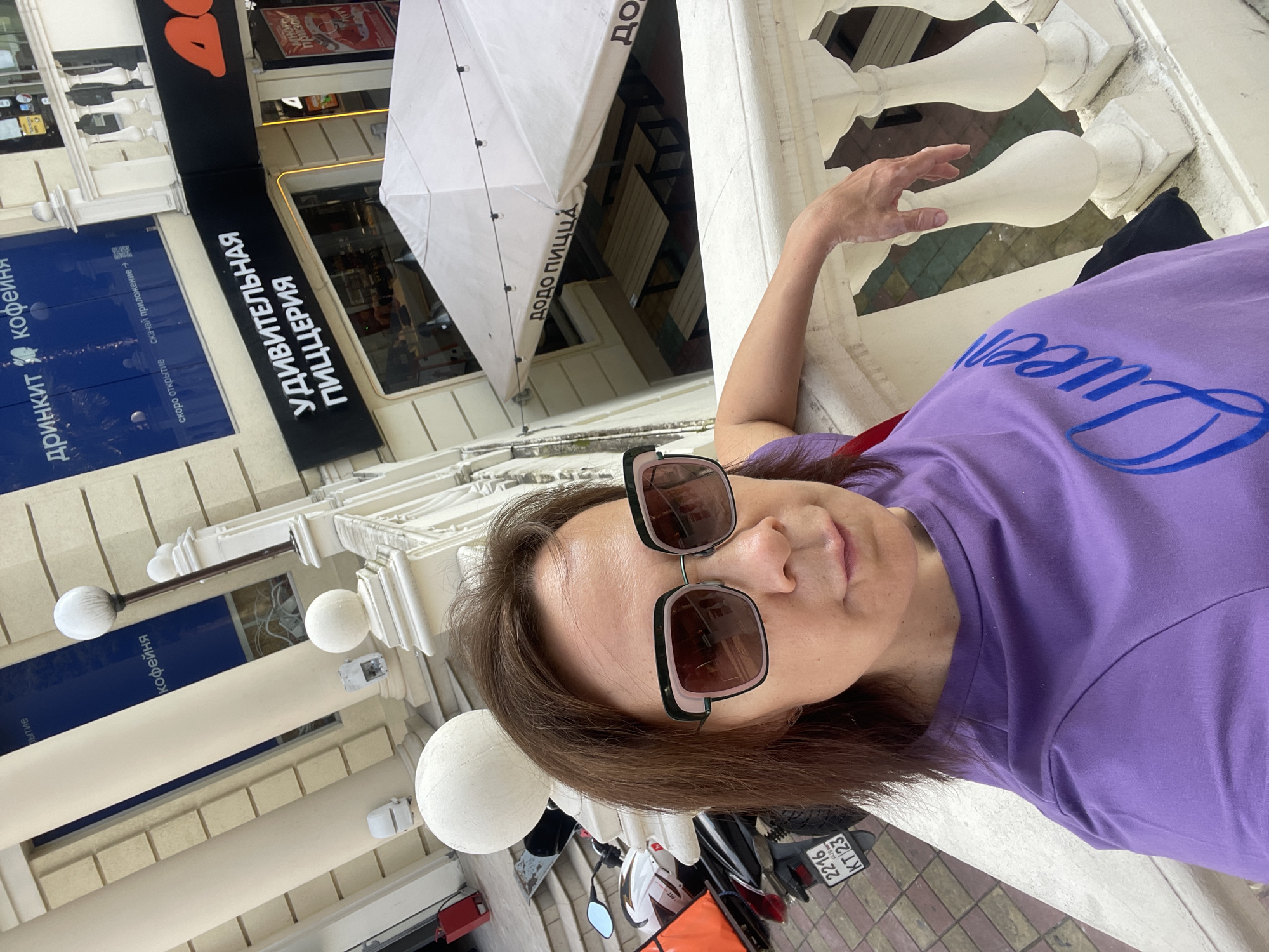 The image shows a woman wearing a purple t-shirt with the word "Queen" printed on it in blue, and oversized sunglasses. She is leaning on a white ornate railing. Behind her, there is a building with several signs. One sign reads "ДРИНКИТ КОФЕЙНЯ" (Drinkit Coffee House) with text below indicating it is opening soon. Another sign reads "УДИВИТЕЛЬНАЯ ПИЦЦЕРИЯ" (Amazing Pizzeria). A white umbrella with the text "ДОДО ПИЦЦА" (Dodo Pizza) is visible in front of the pizzeria. In the background, a motorcycle with a white helmet and an orange bag is parked. The location is identified as Sochi, Russia, based on the text on the motorcycle's license plate which reads "2216 KT 23". The overall scene appears to be an outdoor area of a commercial establishment in Sochi.