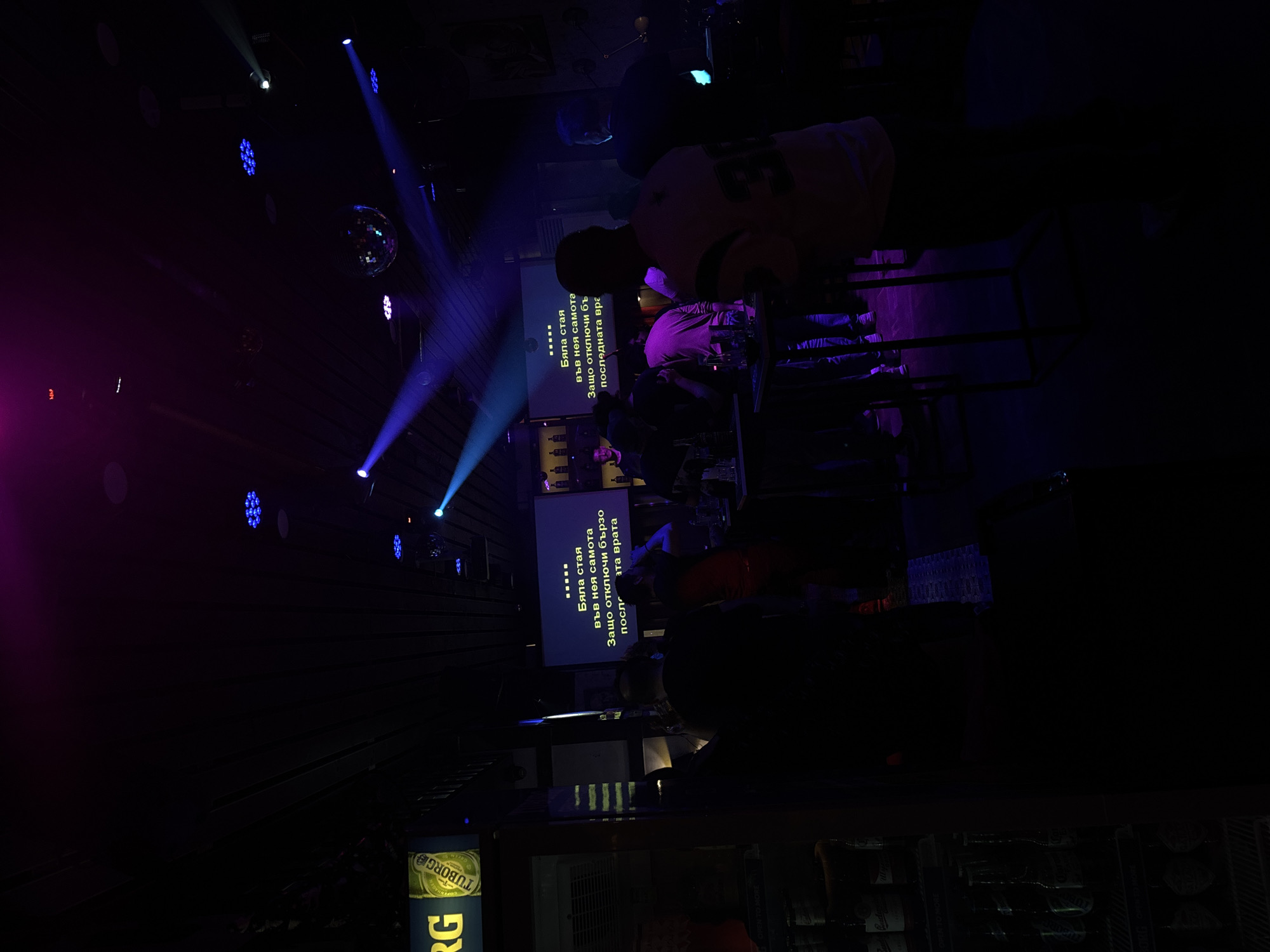 The image depicts a dimly lit interior, likely a karaoke bar or club in Burgas, Bulgaria, based on the Bulgarian Cyrillic script displayed on the karaoke screen, specifically the phrase "Бяла стая във нея самота" and "Защо отключи бързо последната врата," which translates to "White room in it loneliness" and "Why did you unlock quickly the last door."
The scene includes several people seated at tables, mostly obscured by the low light and objects like bottles and glasses. One screen displays karaoke lyrics in Bulgarian, while another possibly does the same. Above the tables, there is a disco ball and lighting fixtures that project blue and purple beams of light. A "TUBORG" branded cooler is partially visible in the foreground. The general atmosphere is one of entertainment or nightlife.