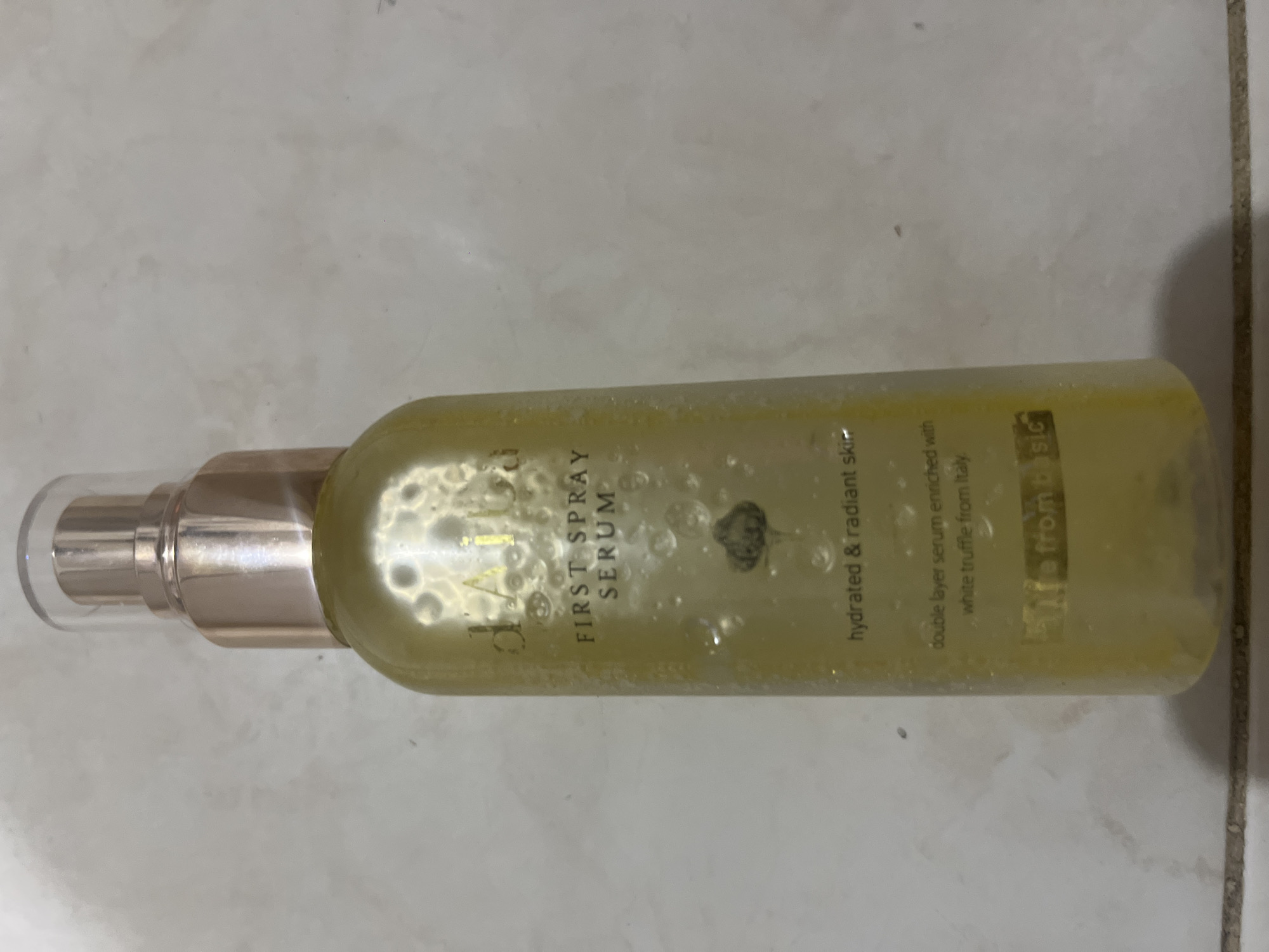 A vertical, close-up shot shows a bottle of facial serum against a tiled background. The bottle, which is primarily clear with a golden liquid, has a rose gold pump and a clear cap. Text on the bottle reads "d'Alba FIRST SPRAY SERUM" and "hydrated & radiant skin double layer serum enriched with white truffle from Italy". The bottle is partially obscured by numerous water droplets on its surface. The background consists of light-colored, rectangular tiles, with some areas appearing slightly darker and more worn, suggesting an indoor setting that may be a bathroom or vanity area. No people or specific landmarks are visible, and there is no information to suggest a location in Denpasar, Indonesia.