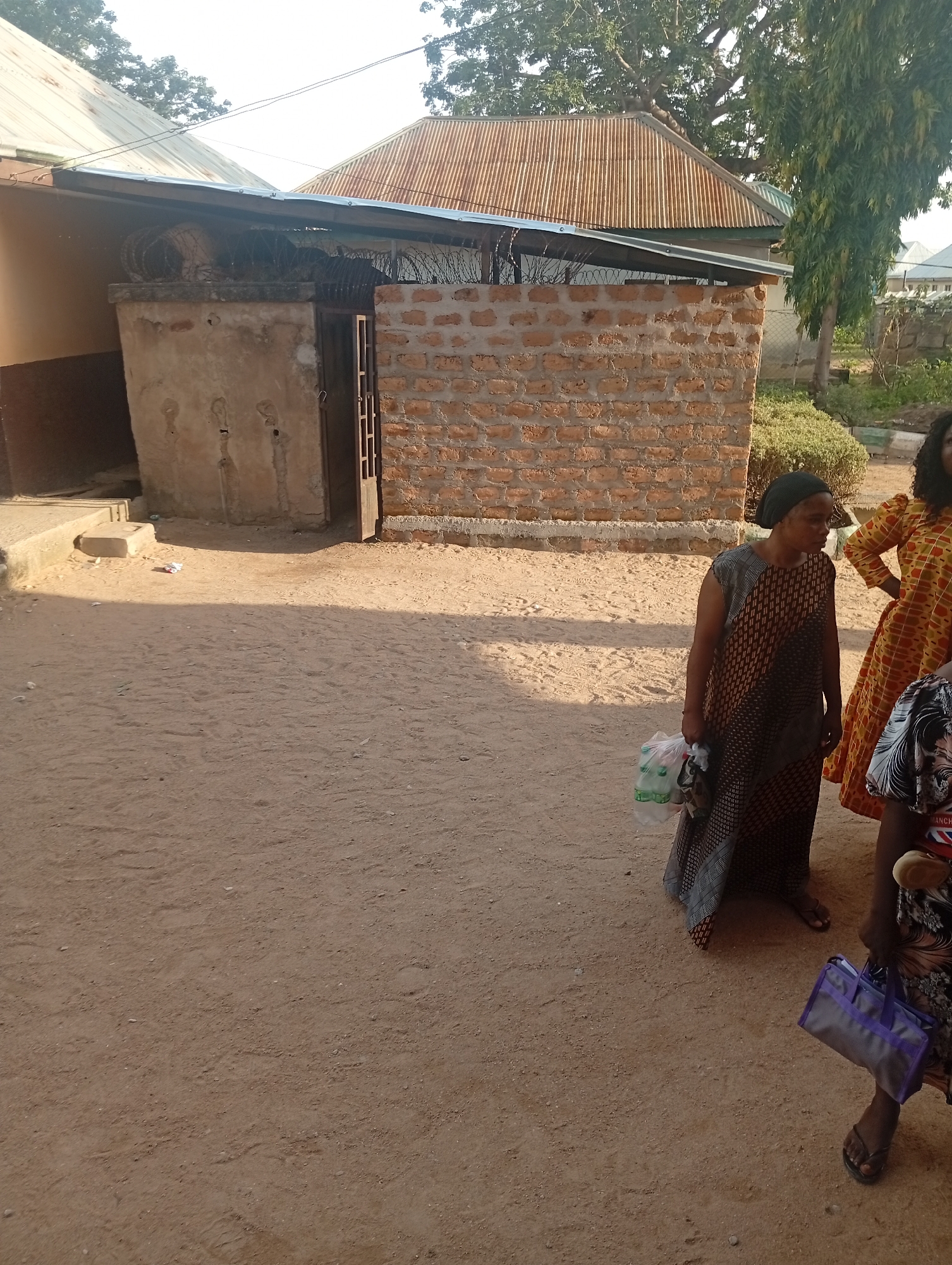 The image depicts an outdoor scene in Jalingo, Nigeria, under what appears to be clear, sunny weather, suggesting it is daytime. The foreground is dominated by a sandy or dirt ground, with some footprints visible. To the right, three individuals are partially visible. One woman in the foreground, wearing a dark headscarf and a patterned dress, holds a plastic bag filled with items. Next to her, another woman is seen wearing a vibrant yellow and orange patterned dress. A third person, also on the right, is partially visible from the waist down, wearing a patterned garment and holding a purple bag.
In the background, a rustic building with a corrugated metal roof is visible. This structure appears to be made of brick and concrete, with some elements of barbed wire visible on top of a wall. A metal gate is also present, partly open, leading into the building. The overall setting suggests a somewhat undeveloped or rural environment. The people's attire and the building's construction are consistent with a location in Nigeria. No specific activity or event is clearly discernible, but the presence of people carrying bags implies they may be going about daily errands or movement. The sunlight casts distinct shadows, further indicating it is daytime.