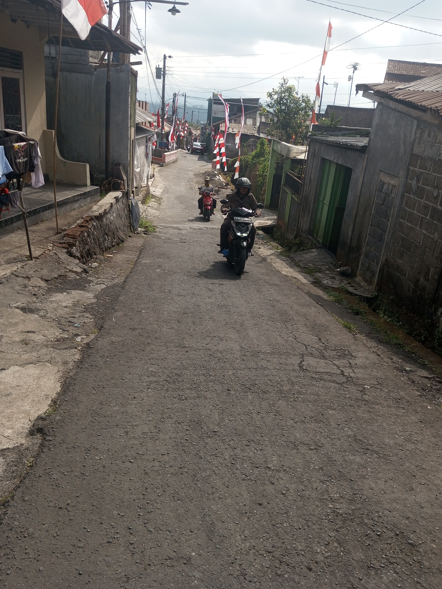 A street in Krasak, Indonesia is depicted in this image, decorated with numerous red and white flags. The street is paved with asphalt and appears to be somewhat aged, with visible cracks. On the left side of the street, there is a building with a porch and laundry hanging outside. The right side features buildings constructed with brick and concrete materials. Several motorcycles are visible on the street; one is in the foreground, ridden by a person wearing a helmet and a brown jacket. Another motorcycle is further up the street, also with a rider. The sky is overcast. The overall impression is that of a residential street decorated for a local celebration or event.
