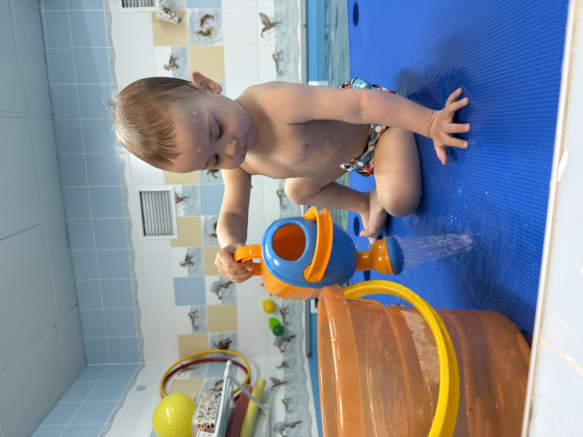 A young child is sitting on a blue textured mat, possibly in a room tiled with blue and white squares. The child is fair-skinned, wearing swim trunks, and has short, wet hair. The child is focused on pouring water from a blue and orange watering can into an orange bucket with a yellow handle. The surrounding area includes tiled walls with pictures of birds. A yellow ball and a hula hoop are also visible. The exact location of the photograph is uncertain and cannot be definitively determined as Rusanovo, Russia.