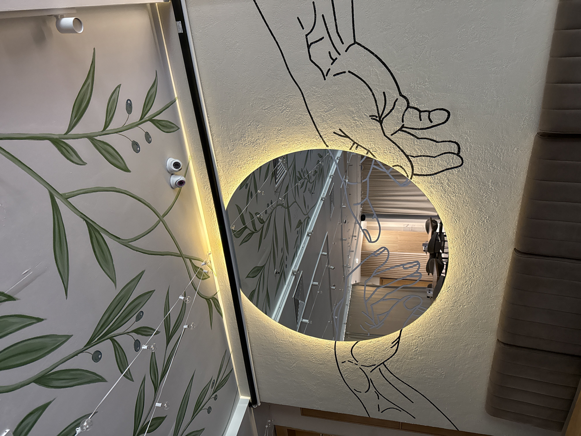 The image captures an interior space in Saint Petersburg, Russia. The walls are decorated with painted olive branches and linear illustrations of hands. A circular mirror with integrated lighting reflects the opposing wall, creating a visual doubling effect of the painted decorations and string lights. The scene is illuminated by the mirror's light, casting a warm glow. A textured couch is visible along the right edge.