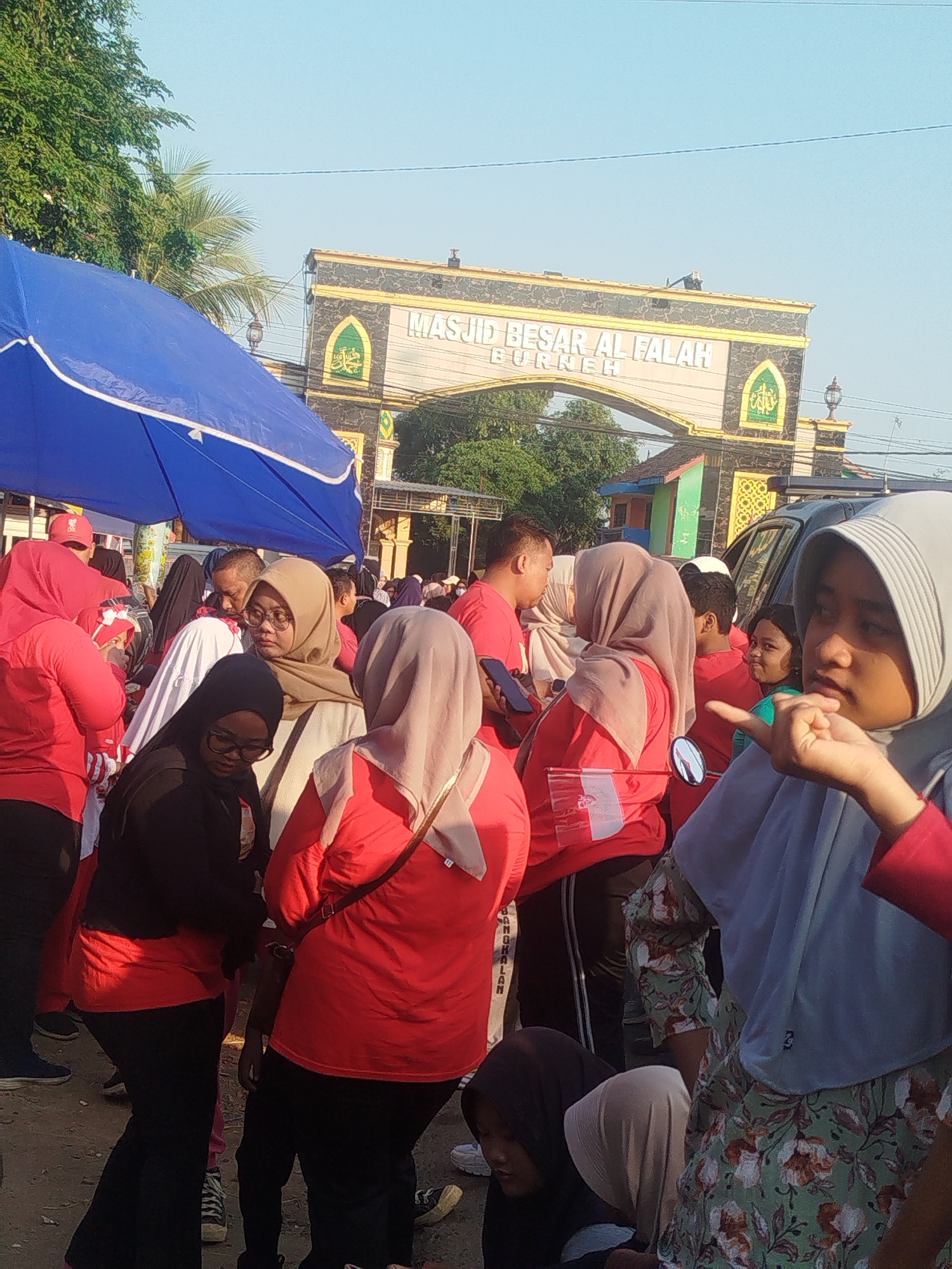 In the city of Burneh, Indonesia, a group of people are gathered in front of an archway with Arabic inscriptions and decorative Islamic motifs. The archway, labeled "MASJID BESAR AL FALAH," appears to be an entrance to a mosque. The area is paved with dirt and gravel, and a blue tarp is set up to one side, possibly for shade or a vendor. Many individuals are wearing red shirts and headscarves, suggesting they are part of a unified group or event. A woman in the foreground, wearing a light blue headscarf and a floral-patterned top, is gesturing with her hand. The lighting suggests it is either early morning or late afternoon, with clear, bright sunlight.