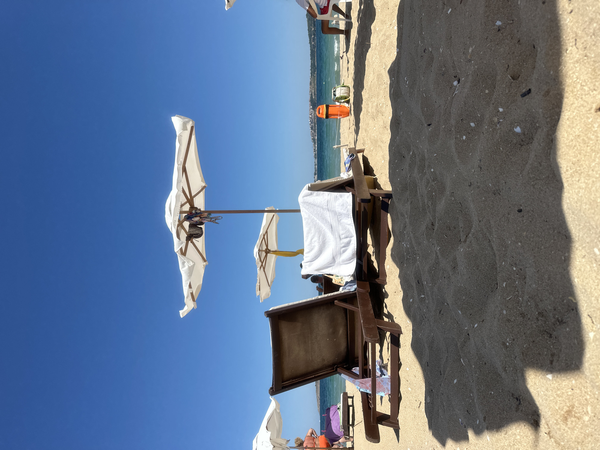 The image depicts a sunny day at a beach, likely in Sozopol, Bulgaria, given the characteristic blue sea and distant coastline with buildings. In the foreground, two empty wooden lounge chairs are positioned on the sand. One chair, closer to the camera, has a white towel draped over its back and a patterned cloth on its seat. The second chair, further back and to the left, has a purple towel and other items placed on it. Both chairs are positioned under large white beach umbrellas.
In the middle ground, the turquoise sea is visible, with gentle waves breaking on the shore. A lifeguard's orange buoy and rope are placed on the sand near the water's edge. Further out, the coastline of Sozopol is visible, with buildings dotting the landscape. Two people are partially visible in the background. One person, wearing an orange swimsuit, is standing near a lounge chair on the left side of the frame, while another person in a white shirt and red shorts is seated in a white chair on the right. The sky is a clear, vibrant blue. The sand in the foreground is light brown and textured, with small shells and debris scattered across it. Shadows from the umbrellas and chairs are cast on the sand, indicating the sun's high position in the sky.