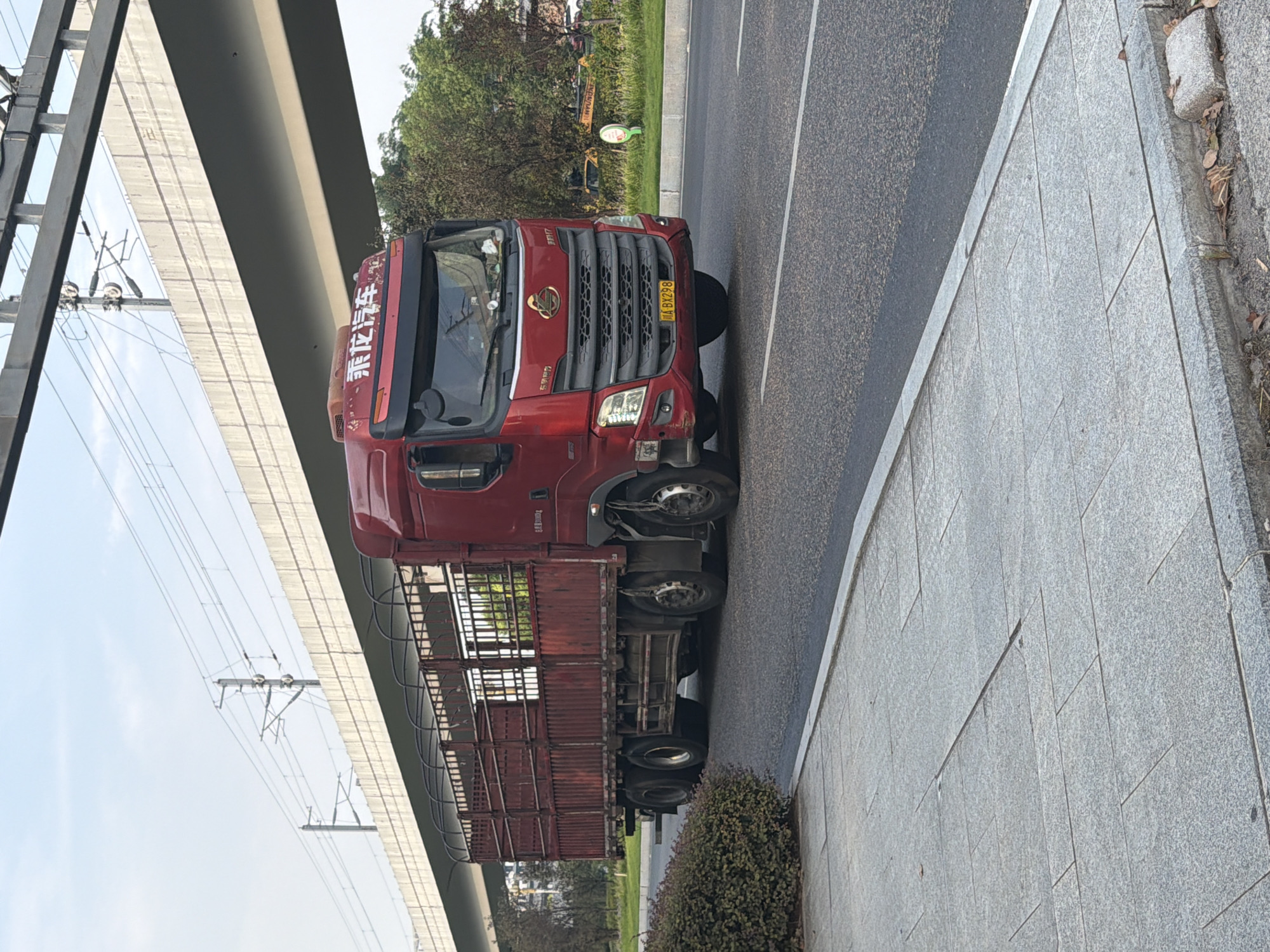 A red truck with a cage trailer drives on a road under an elevated highway in Jiangjin, China. The truck has a license plate that reads "WA BX298". The sky is clear and blue. Trees and buildings are visible in the background. The road is paved with asphalt, and there is a sidewalk made of gray stone slabs. The truck appears to be moving forward.