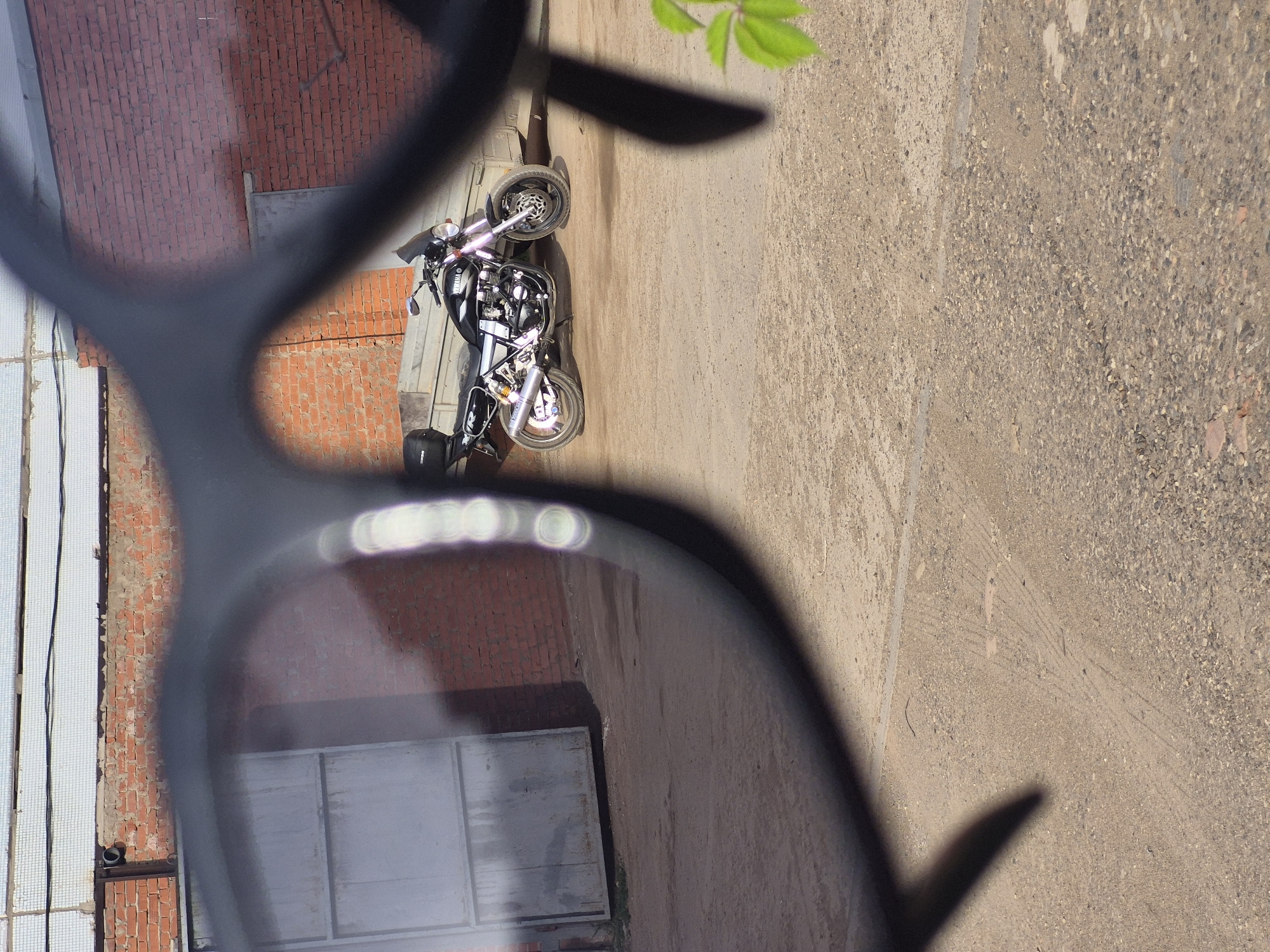 A street scene is visible through the lens of sunglasses. A motorcycle is parked near a red brick building, next to a metal door. The ground is concrete. A green leaf is visible at the top of the frame.