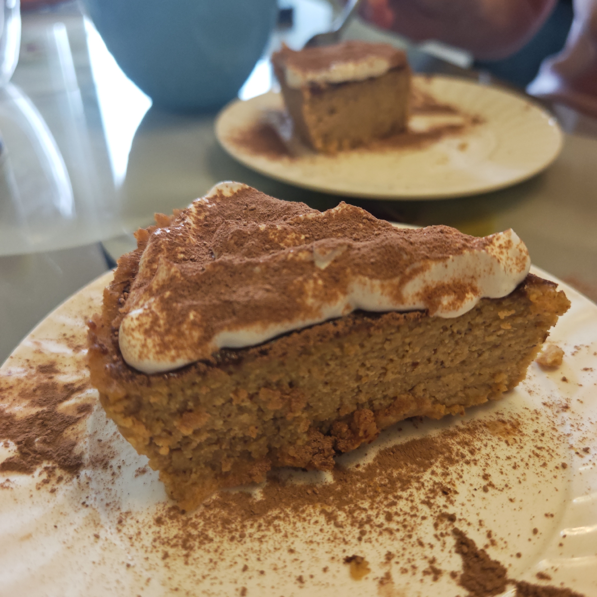 The image shows two plates of what appears to be pumpkin pie or cake, topped with white cream and dusted with a brown powder (possibly cinnamon or cocoa). The plates are white. The larger slice is in the foreground and is cut at an angle. The smaller slice is in the background. Both plates have the brown powder scattered around the slices. The background also includes a blue cup or bowl and blurred figures that may be people. The setting appears to be indoors on a light-colored table. It is not possible to discern the location or city of Rostov, Russia, from the image.