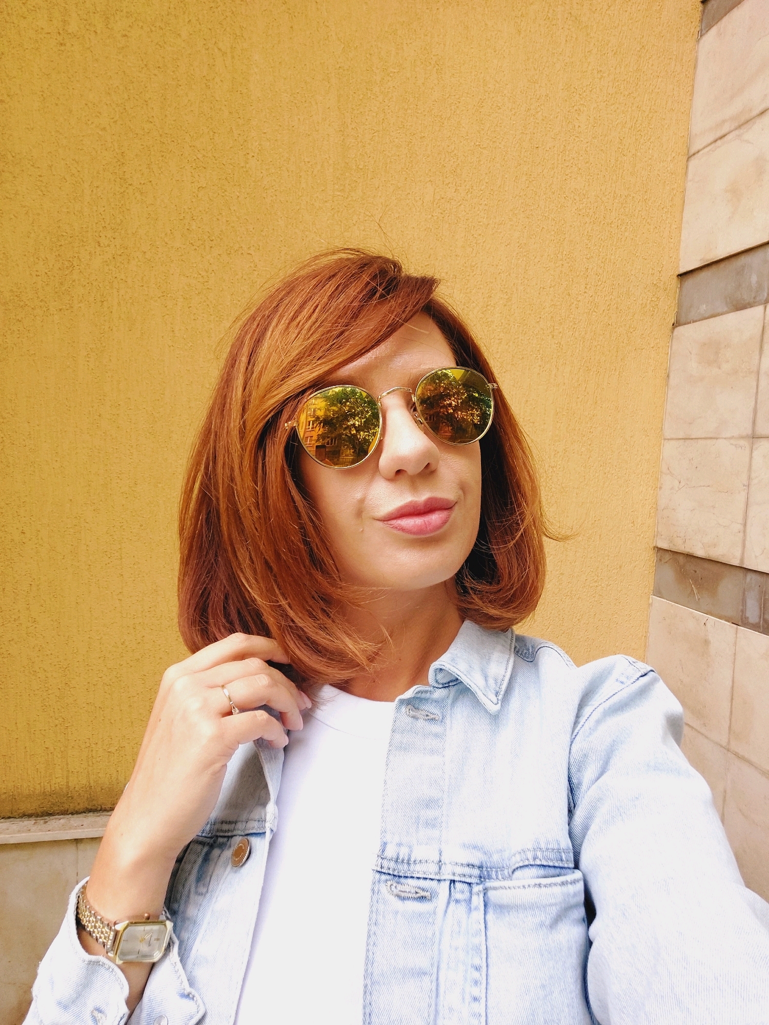 A woman with auburn hair and sunglasses stands in front of a textured yellow wall with a tiled vertical structure to her right. She is wearing a light blue denim jacket over a white t-shirt, a gold watch on her left wrist, and a ring on her left ring finger. The sunglasses have round, reflective lenses. The scene is outdoors, possibly on a street or patio, in a city. The backdrop suggests an urban environment, but no specific landmarks in Sofia, Bulgaria, are identifiable from the image alone.