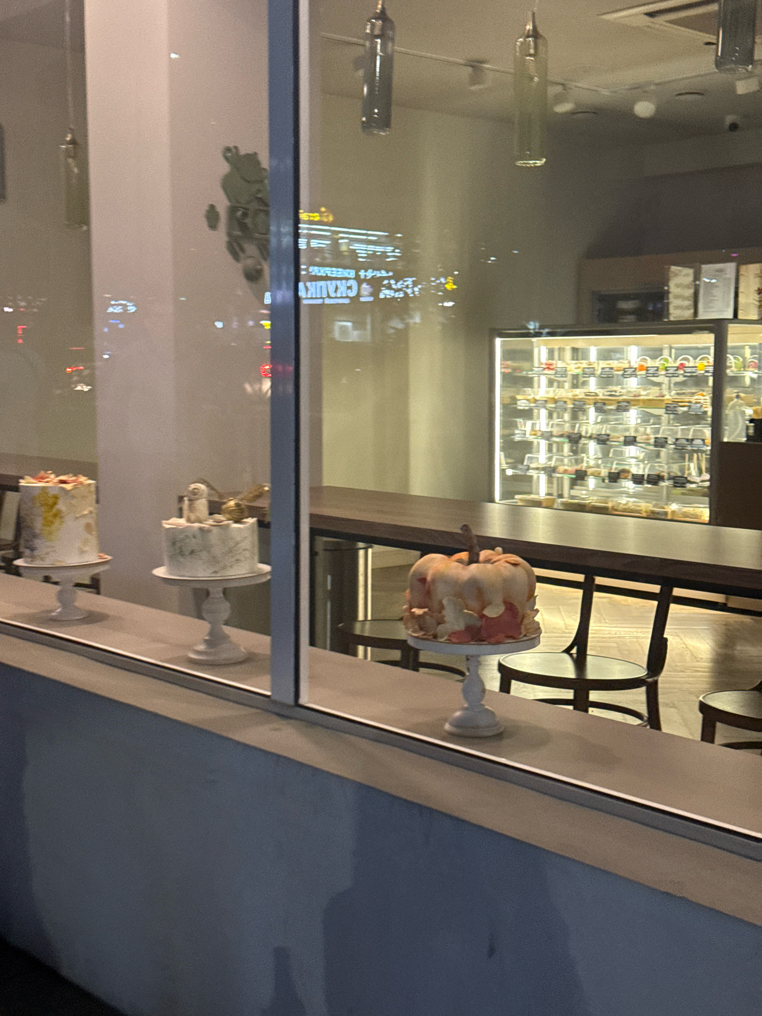 The image shows a view through a window into what appears to be a bakery or cafe. Inside, three decorated cakes are displayed on white pedestals on a counter. The cake on the left is a round, white cake with gold and pink accents. The middle cake is also round and white, decorated with green marbling and topped with a white figure and a gold snitch. The cake on the right is shaped like a pumpkin with pink and orange frosting dripping down.
Behind the counter, a refrigerated display case is filled with rows of individual pastries or desserts. The interior of the establishment appears modern, with a long wooden table and chairs visible in the background. Reflections on the window show lights and possibly street traffic from outside, suggesting it is evening or night. The overall setting is clean and well-lit.