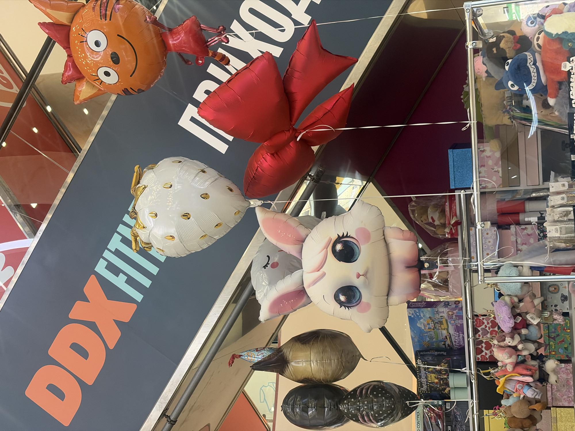 The image depicts an interior setting in Saint Petersburg, Russia, showcasing a variety of balloons and merchandise. In the foreground, a collection of helium balloons featuring characters like an orange tiger-like figure, a red bow, a white spotted balloon, and a cartoonish rabbit hang suspended. The balloons are connected by strings and appear to be for sale or decoration. Behind the balloons, a sign with the inscription "DDX FITNESS" is visible. To the right, a display case is stocked with various items, including stuffed animals, toys, and gift boxes. The background shows a shopping mall interior with multiple levels and escalators.