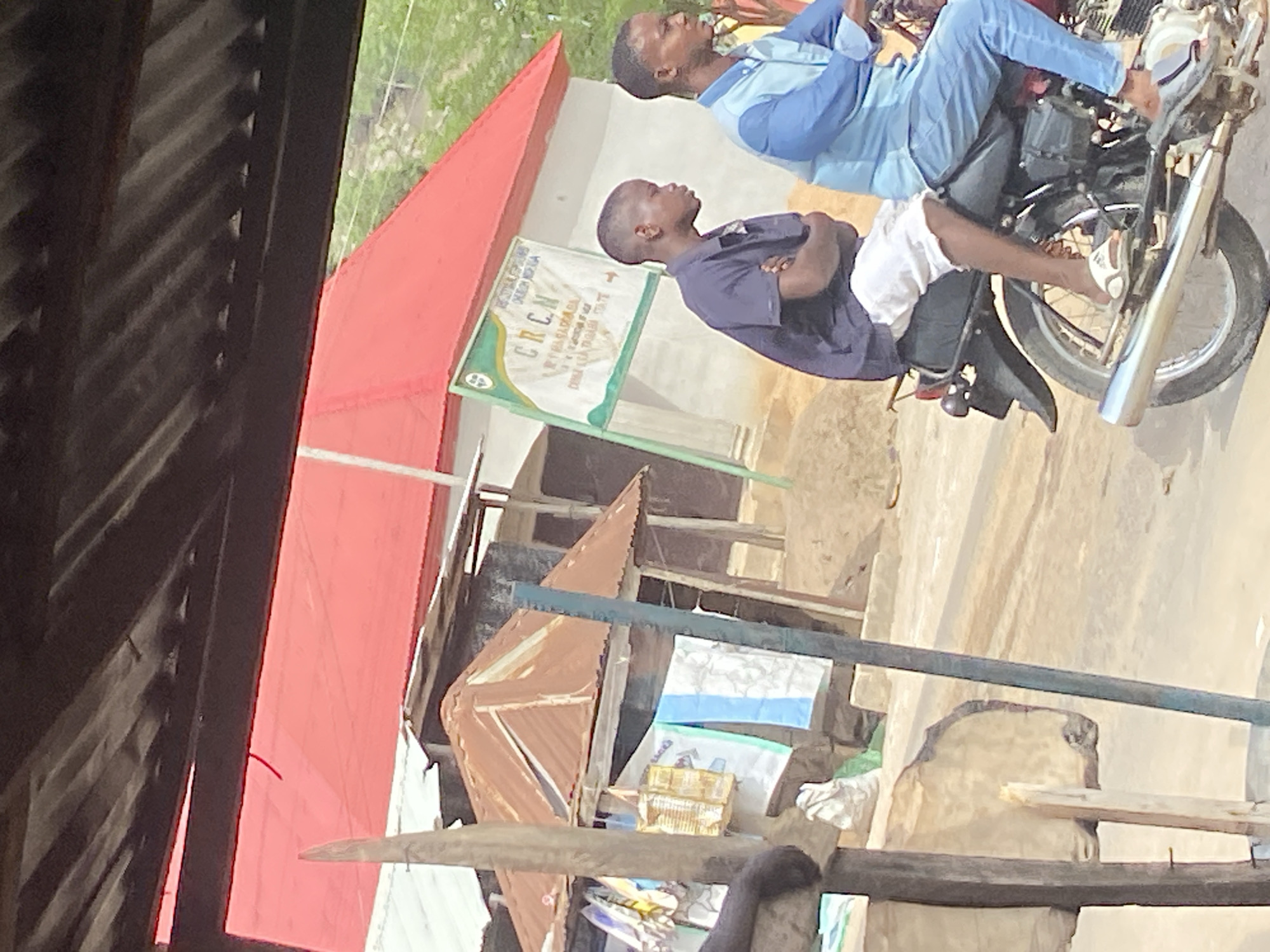 The image captures an outdoor scene in Mararaba, Nigeria, depicting what appears to be a street or communal area, viewed from within a shaded structure. Two young men are central to the photograph, both seated on a dark-colored motorcycle. The man in the foreground is dressed in a navy blue t-shirt and white shorts, while the man behind him wears a light blue long-sleeved tunic and dark blue trousers. Both individuals are looking towards the left side of the frame, with neutral expressions, suggesting they are either waiting or observing.
In the midground, a building with a distinctive bright red corrugated iron roof is prominent. Attached to a wall near this building is a green and white sign. Visible text on the sign includes "KOTALAN KWAN", the prominent abbreviation "CRCN", and other phrases such as "SADAR DA KOWA" and "YUNIYA 2017". The immediate foreground shows dark wooden slats and beams, indicating the photograph was taken from inside a simple structure or stall. Other makeshift buildings or fences constructed from corrugated metal sheets are also visible.
The scene is illuminated by bright daylight, suggesting sunny weather. The overall environment portrays a functional and somewhat informal setting, characteristic of the region.