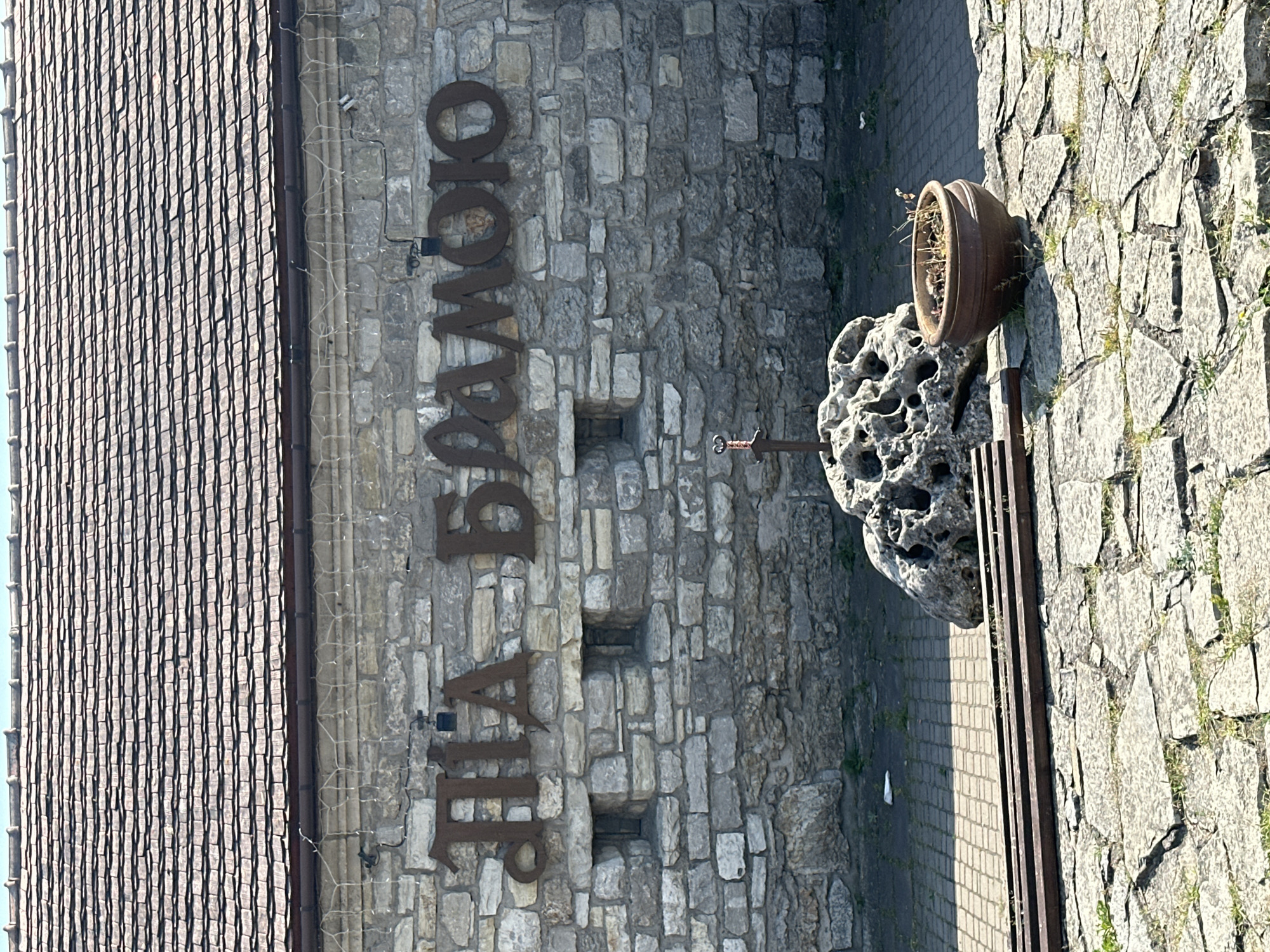 This is a photograph of the stone wall of a building in Kamianets-Podilskyi, Ukraine, featuring large, rusty metal letters spelling out "ПІА БЛАМОЮ" (PIA BLAMOJU) above two small, recessed windows. In front of the wall, a weathered, porous rock formation stands with a decorative sword embedded in its top. To the right of the rock, a brown ceramic pot contains dry plants. The ground in front of the wall is paved with small, patterned bricks, and a portion of the wall is constructed from rough-hewn stones. A section of metal shelving or track is visible in the foreground, and the roof of the building, made of dark tiles, is visible at the top of the image.
