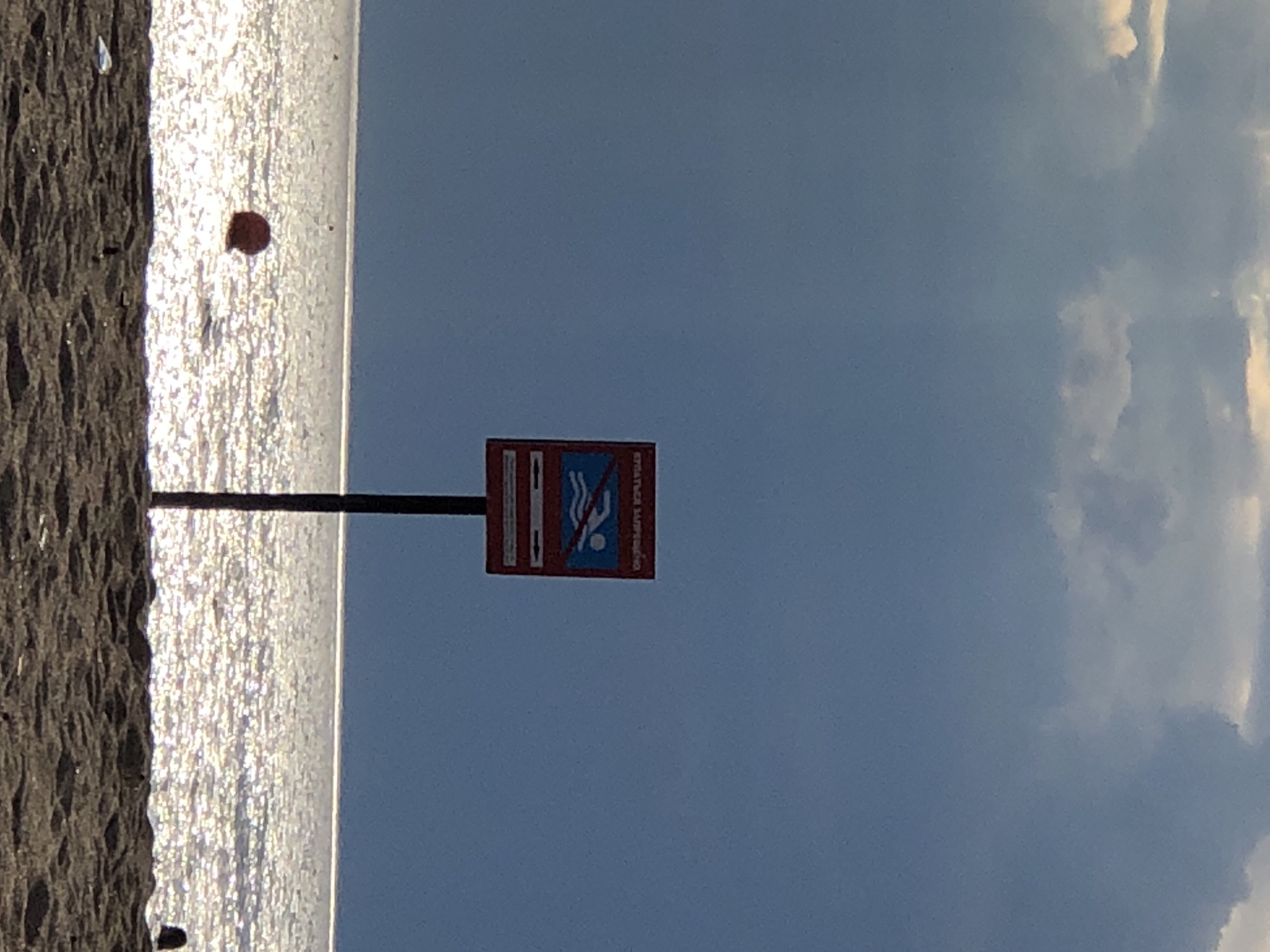 The image captures a beach scene in Sestroretsk, Russia, under a dramatic sky. The upper portion of the frame is dominated by heavy, dark blue-grey clouds, with some lighter patches indicating partial illumination. The horizon line is visible as a bright, thin strip where the dark sky meets the highly reflective sea.
The sea surface shimmers intensely with glints of light, creating a sparkling effect across the water. A single, distinct red spherical buoy floats in the water to the left of the image's center.
In the foreground, a dark sandy beach with visible undulations and footprints extends across the lower part of the frame. A small piece of white debris is discernible on the sand in the lower-left corner. On the far right edge of the beach, a dark, indistinct rounded shape, possibly a person's head or a small object, is partially visible.
Centrally positioned on the beach, a dark pole supports a rectangular red sign. The sign bears Cyrillic text at the top, which translates to "SWIMMING IS FORBIDDEN". Below the text, a graphic features a white swimmer symbol on a blue background, traversed by a red diagonal line, explicitly prohibiting swimming. Below this symbol, two white horizontal arrows point left and right on a blue background, likely indicating the boundaries of the forbidden area or directing people.