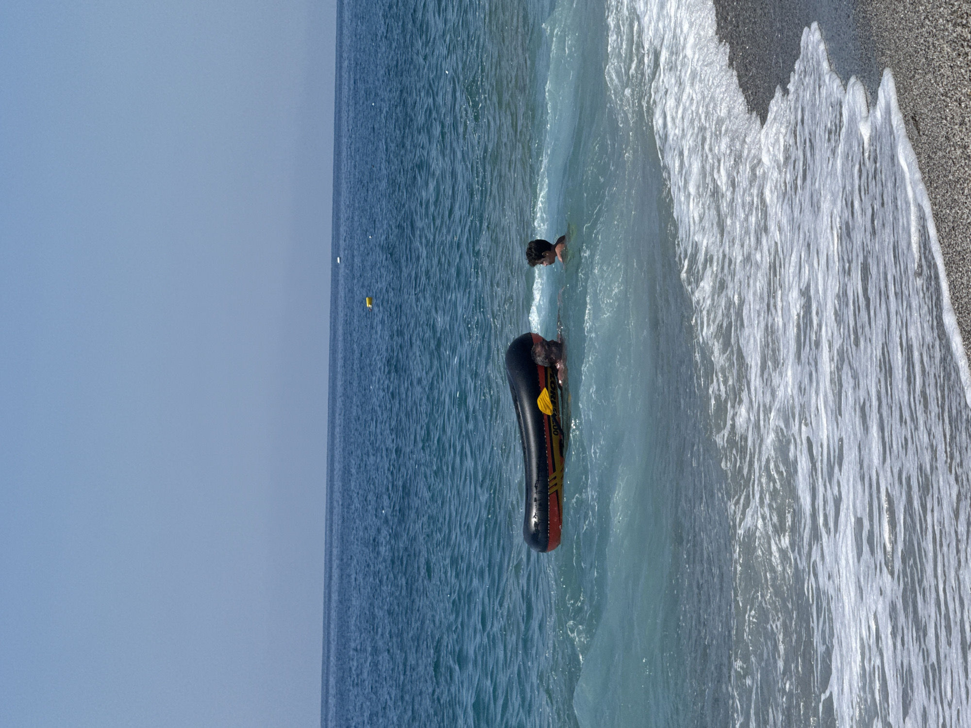 Two individuals, one adult and one child, are in the sea near the shore of Alanya, Turkey, playing with an inflatable raft. The adult appears to be partially submerged in the water behind the raft, while the child is in front of it, facing away from the viewer. The raft is black and red with yellow accents. The water is a clear blue-green, with waves breaking on a pebble beach in the foreground. The sky is a clear, pale blue. There are a few yellow buoys visible in the distance. The overall scene depicts a recreational activity at a beach.