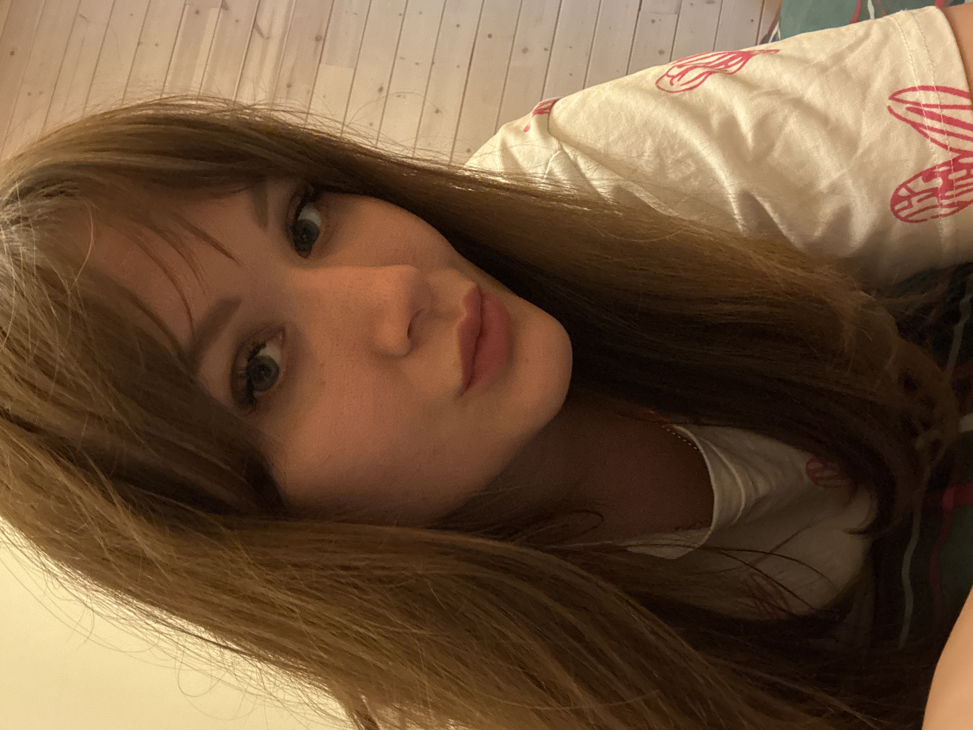 A close-up selfie shows a young woman with long, light brown hair parted to the side. She is wearing a light-colored t-shirt with pink butterfly wing prints. Her face is angled towards the camera, with her lips pursed in a kiss. The background includes a light-colored wooden floor and hints of a patterned surface that could be bedding or fabric. No specific location is discernible in Taman', Russia.