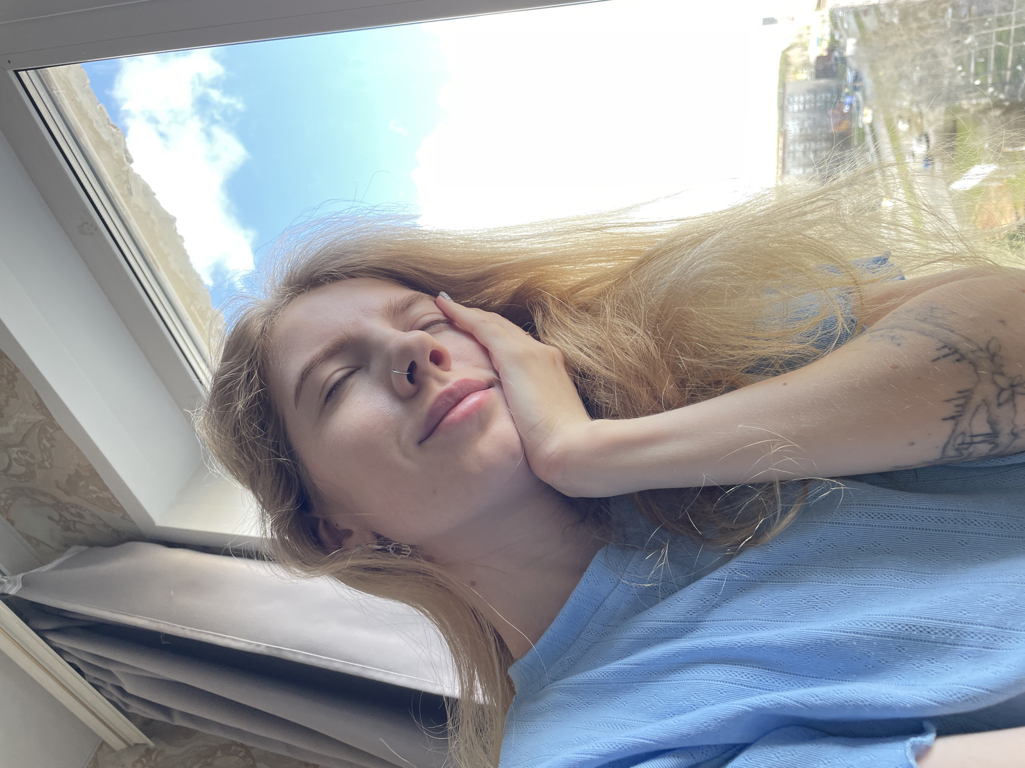 A young woman with light blonde hair and fair skin is captured in a close-up, upward-angled shot. Her eyes are gently closed or squinting, and she has a small nose ring. She rests her left hand on her cheek, appearing peaceful and relaxed. She wears a light blue, textured long-sleeved top, and a tattoo, possibly depicting figures or a scene, is visible on her right forearm.
The setting places her near a white window frame, which reveals a bright blue sky with scattered white clouds outside. The strong natural light illuminates her hair and face. Through the window, to the right, blurred outlines of buildings are visible, suggesting an urban or suburban environment, aligning with the provided context of Izhevsk, Russia. To her left, part of a wall with patterned wallpaper and grey/beige curtains can be seen, indicating an indoor setting. The image conveys a sense of calm and enjoyment of a sunny daytime, with clear skies. No visible text is present.