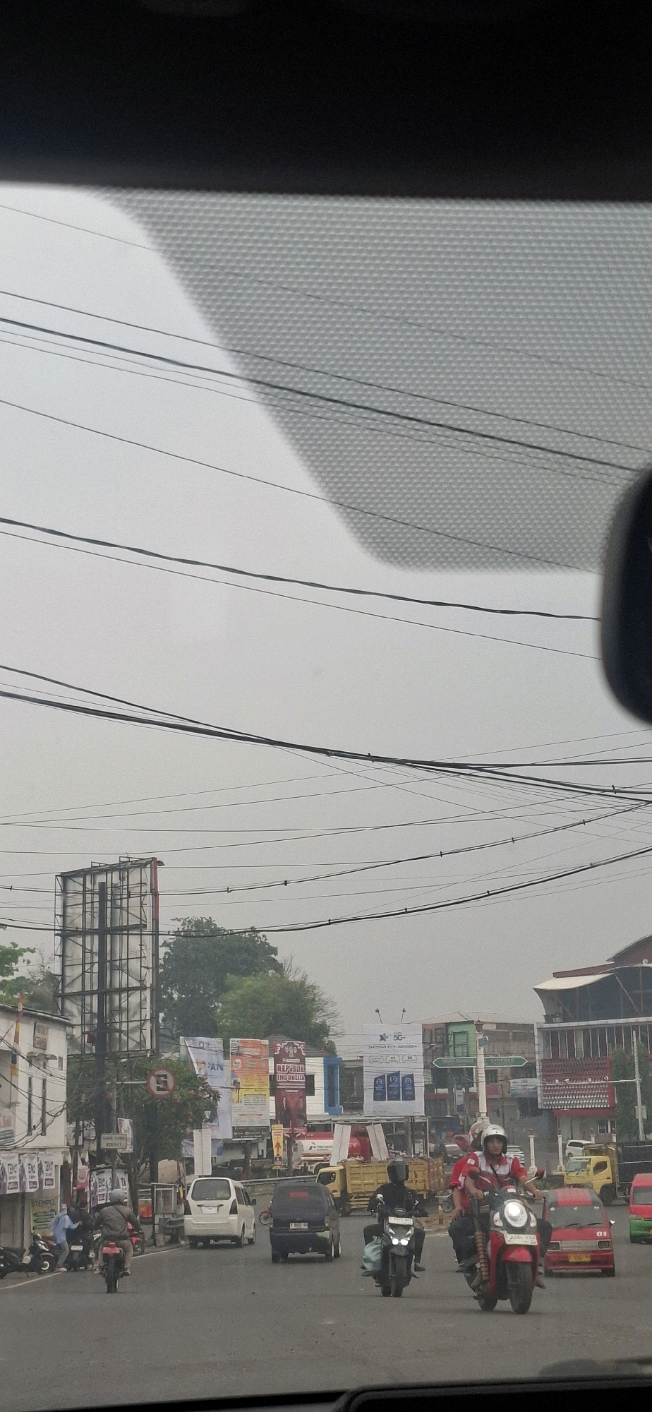 The image shows a street scene in Sirnagalih, Indonesia, on a hazy day. The perspective is from inside a vehicle, looking forward through the windshield. The sky is overcast and a network of power lines crisscrosses the upper portion of the frame. In the mid-ground, there is a street with several vehicles including motorcycles, cars, and a yellow truck. On the left side of the street, there are buildings and a large billboard. Various advertisements are displayed on banners and billboards along the street. People are visible on motorcycles, some wearing helmets. The general atmosphere suggests a busy urban environment.