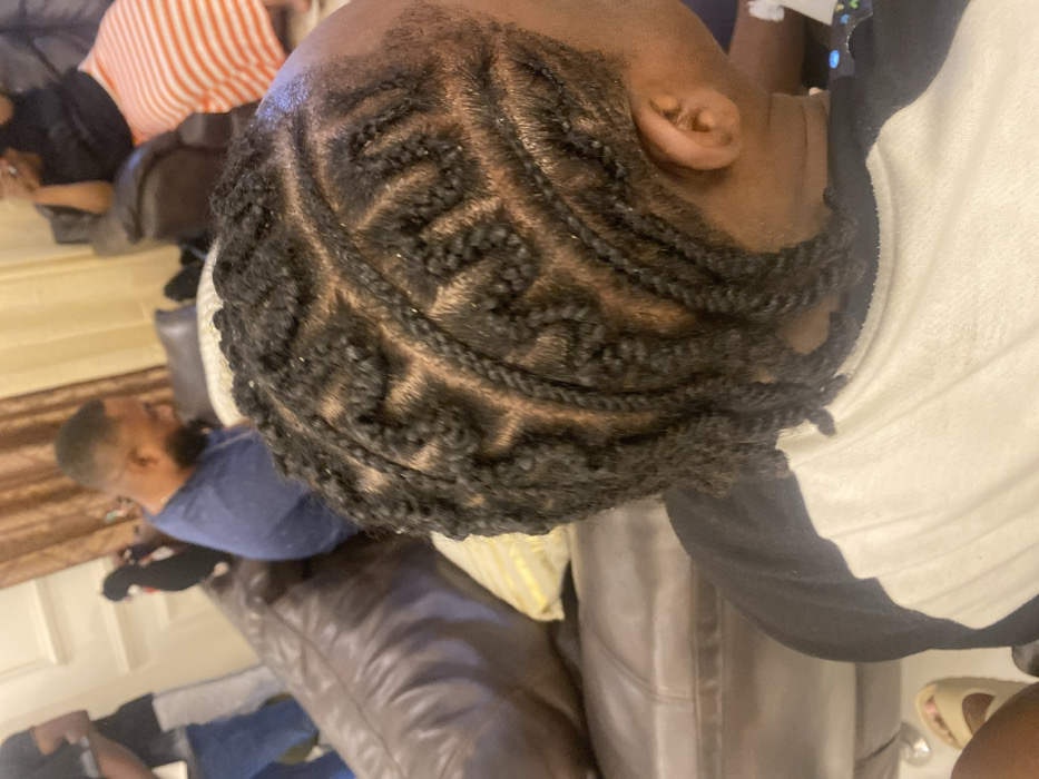 This image, set in Adenta Municipality, Ghana, provides an overhead view of an indoor scene, likely a living room. The primary subject is a person whose head is prominently featured, showcasing an intricate hairstyle of dark, tightly braided cornrows with defined geometric patterns. Their right ear is visible, and they are wearing a white top.
In the background, several other individuals are partially visible. To the mid-left, a person wearing a dark blue top is seated on a large brown leather couch, facing away from the viewer. Further back on the left, another individual is seen wearing an orange and white striped top. In the bottom right corner, a leg and a foot wearing sandals are visible, indicating the presence of a fourth person.
The setting is furnished with multiple brown leather couches, suggesting a comfortable and communal space. White wall paneling and brown window coverings (curtains or blinds) are also discernible. A yellow or gold-colored cushion is visible near the main subject. The lighting suggests it is daytime. The overall atmosphere appears casual and relaxed. No text is visible in the image.