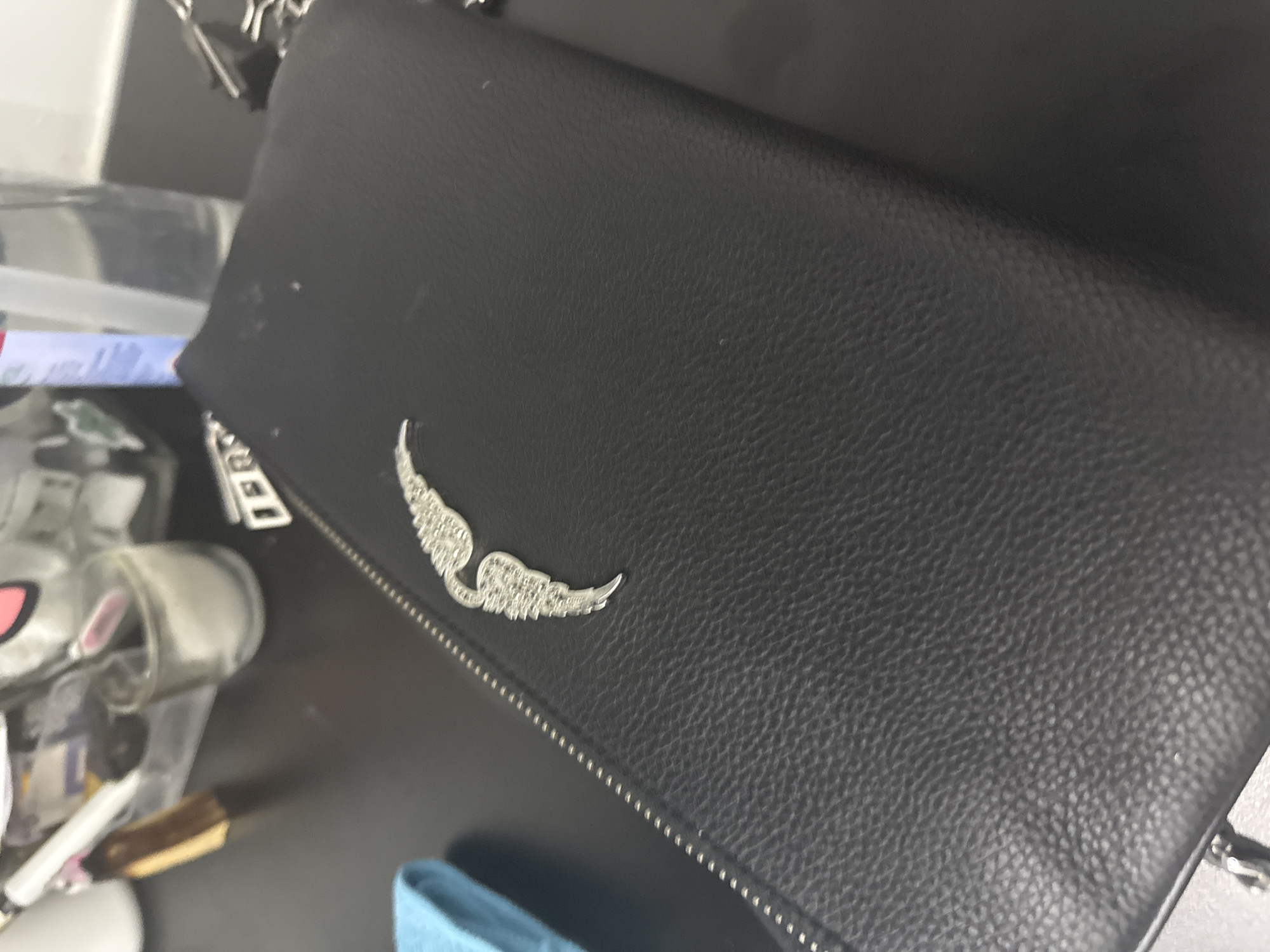 The image displays a close-up view of a black, textured, rectangular object, likely a clutch or wallet, positioned diagonally across a dark surface, possibly a desk, in San Bernardino Tlaxcalancingo, Mexico. A prominent silver-toned metallic emblem, shaped like stylized wings adorned with small, reflective elements, is affixed to the upper left quadrant of the object's visible surface. A metallic zipper runs along the left edge of the black item. To the immediate left, a portion of a light blue cloth object is visible. The upper background, out of focus, reveals several items on the dark surface: a clear container holding various personal effects, including a brush with a brown handle; a small glass containing a white granular substance; a clear bottle, likely containing liquid, with a white cap; and obscured metallic objects, possibly keys or hardware, on the far right. No individuals are present in the scene, which appears to be an indoor setting.