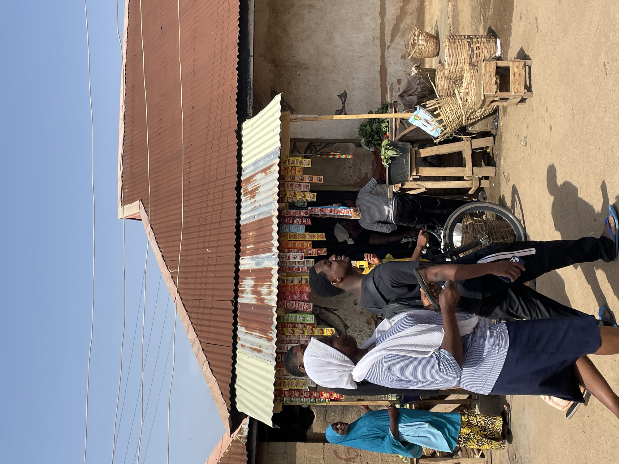 This image captures a vibrant street market scene in Jos, Nigeria, under a bright, clear blue sky. The setting features a rustic building with a rusty, corrugated metal roof and an unpaved, dusty ground. Overhead, several electrical wires stretch across the sky.
At the center of the image is a bustling street stall made of wooden planks, serving as a display for various goods. The stall is laden with numerous small, colorful packaged items, including what appear to be instant noodles (like "INDOMIE"), sweets, and other provisions, arranged on shelves. Green leafy vegetables are also visible on the counter. Beside the stall, woven baskets and wooden crates are stacked, some used for display. A bicycle with a prominent wheel is parked near the stall.
Several people are gathered around the stall. In the foreground, a woman wearing a blue and white striped shirt, a dark skirt, and a white head covering is holding and looking at a smartphone or tablet. Behind her, another person in a black t-shirt and black pants also appears to be focused on a mobile device. Further to the left and back, a person clad in a light blue garment looks upwards. Another individual is partially visible sitting behind the market counter. The bright daylight casts distinct shadows, indicating a sunny day. While specific product names are largely illegible due to resolution and size, the packaging clearly shows that various consumer goods are being sold. The scene depicts a typical daily commerce and social interaction in a local market environment.