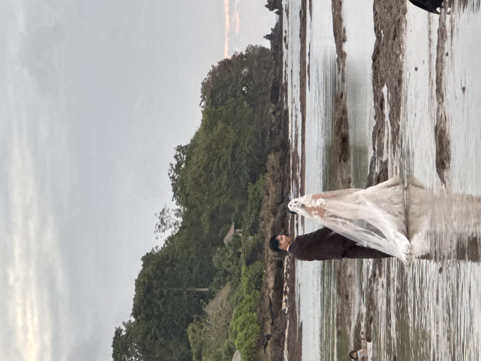 A wedding couple, a man in a dark suit and a woman in a white wedding gown and veil, stand in shallow water on a rocky shore. The couple is facing away from the camera, looking towards a treed cliff face. In the distance, a person can be seen wading in the water. The sky is overcast, and the water reflects the sky. The scene appears to be at the beach in Kuta Selatan, Indonesia.