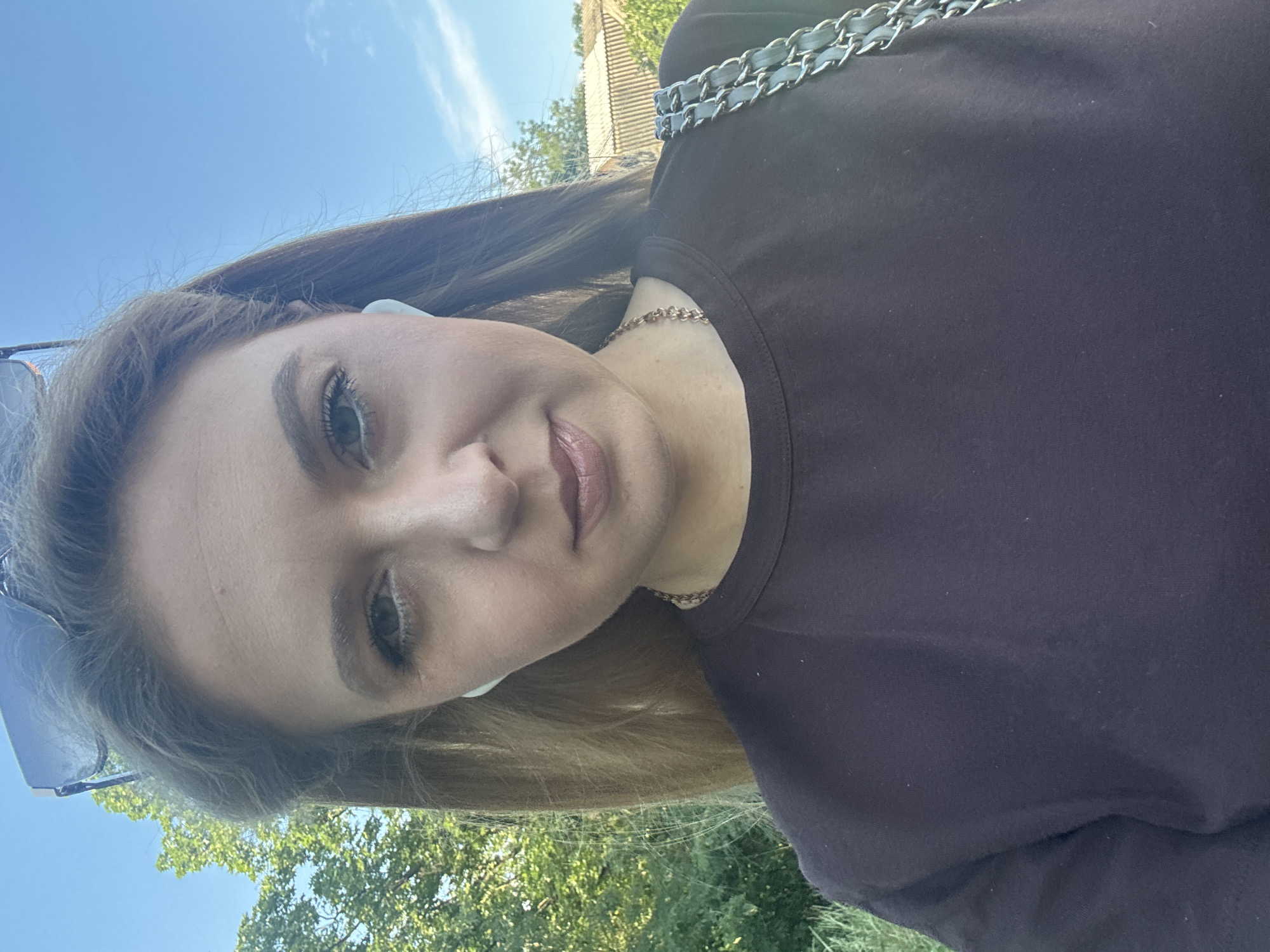 In this close-up, outdoor selfie, a young woman with long, light brown hair is captured from the chest up, against a backdrop of a clear blue sky and lush green trees. She is wearing a dark maroon crew-neck t-shirt and a rose gold chain necklace. A light blue earbud is visible in her right ear, and sunglasses are perched on top of her head. Her left shoulder is adorned with a quilted purse strap featuring silver chain links. The lighting suggests a sunny day, likely late morning or early afternoon, casting a natural glow on her face as she looks directly at the camera with a neutral expression. The setting appears to be a park or a residential area with greenery and a hint of a building in the background, suggesting an urban or suburban environment.