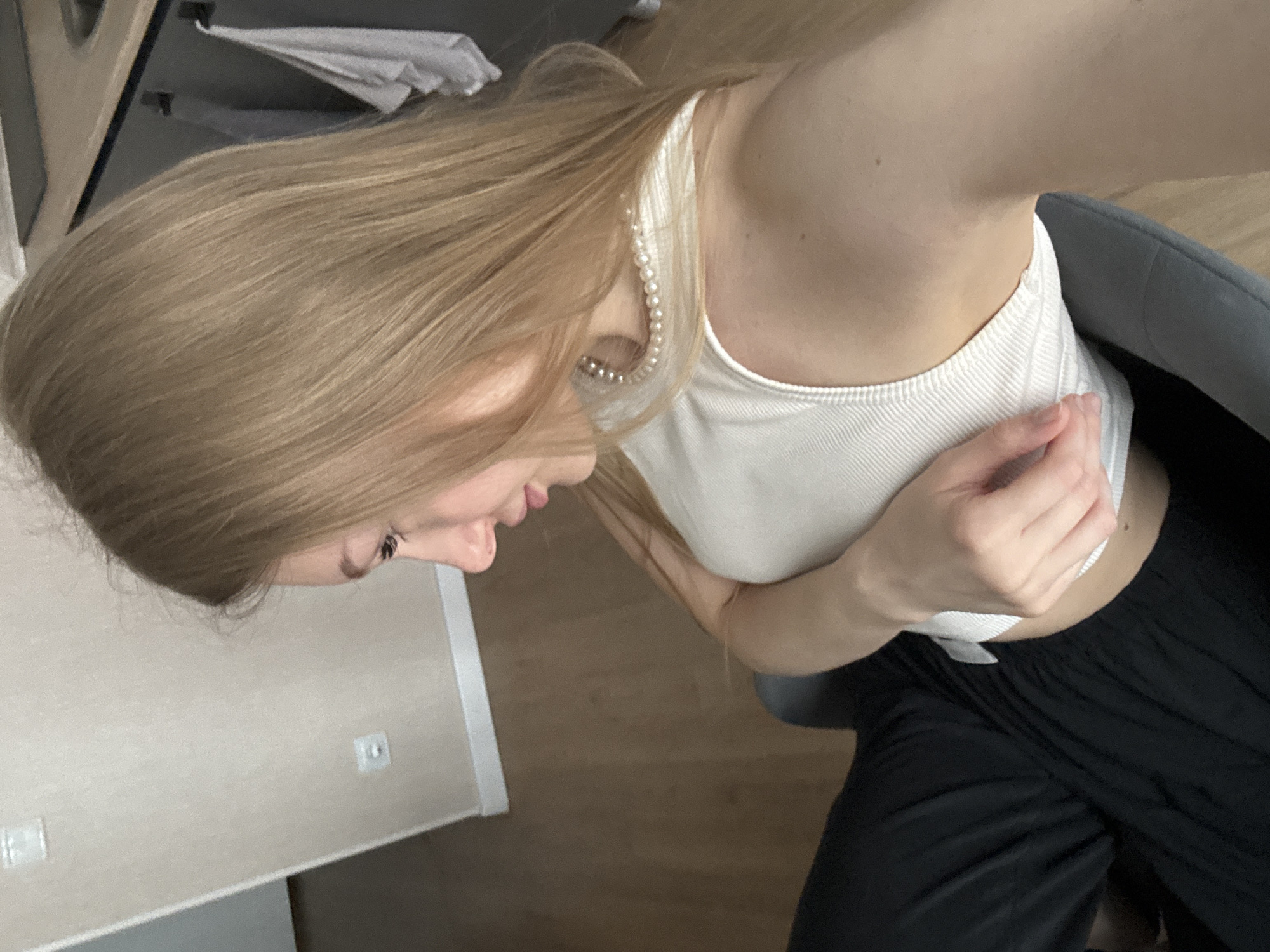 The image, captured in Perm, Russia, depicts a single female individual in an indoor setting. The subject, possessing light skin and long, straight blonde hair, is shown in right profile, seated, and looking downward. She is attired in a white ribbed tank top, a pearl necklace, and black pants. Her right hand is positioned on her midsection, lightly touching the edge of her top. The background features light beige walls with two visible electrical outlets or switches. The floor is light brown wood. To the right, a kitchen area is visible, including a dark countertop, a partially obscured sink, dark cabinetry, and two towels hanging from dark hooks. The subject is seated in a dark grey chair.