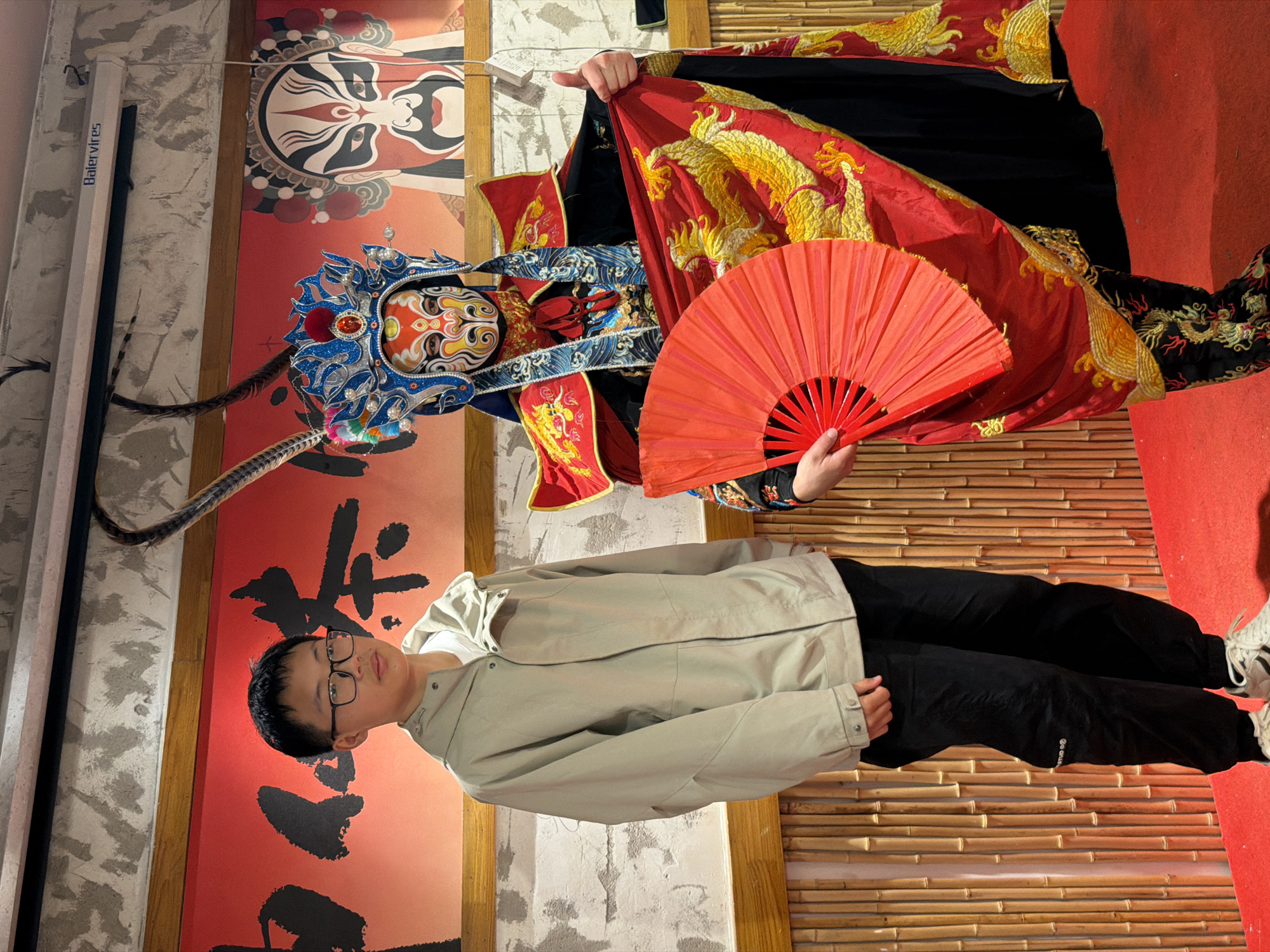 In the city of Yuzhong District, China, a young boy wearing glasses and a light-colored jacket stands to the left of a performer in traditional Chinese opera attire. The performer, with an elaborately painted mask and headdress, holds a red fan and a richly embroidered red garment. The backdrop features a red wall with bold Chinese characters and an image of a painted opera face. The scene is set on a red carpet in front of a bamboo-like textured wall.