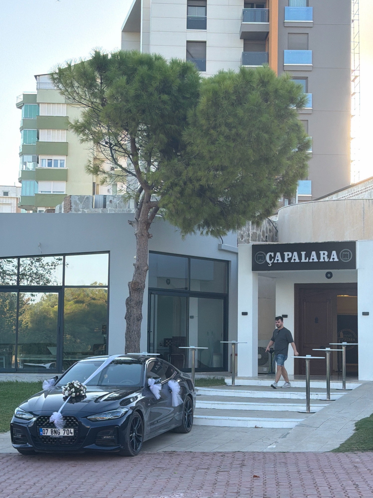 In Antalya, Turkey, a black BMW is decorated with white ribbons and a flower bouquet on the hood, suggesting it is prepared for a wedding. It is parked in front of a modern building with large glass windows, reflecting the surrounding trees and sky. A sign above the entrance reads "ÇAPALARA". A man in a dark t-shirt and denim shorts walks up the steps towards the building. In the background, several tall, multi-story buildings are visible, characteristic of an urban environment. The weather appears to be clear and sunny, with the light indicating it is likely late afternoon.