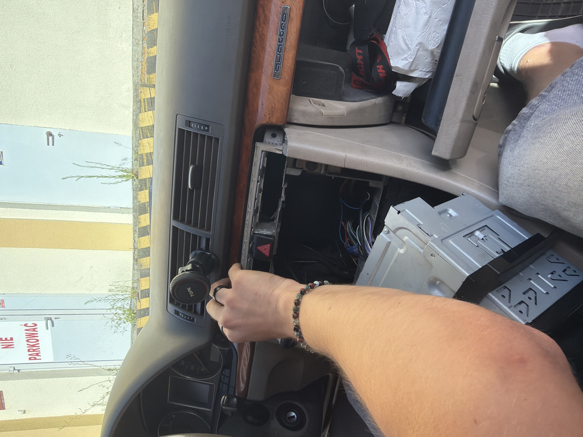 The image shows the interior of a car, likely an Audi, with the dashboard disassembled. A person's arm and hand are visible, reaching into the dashboard area where the car's stereo is being removed or installed. The person is wearing a bracelet. Wires are exposed in the open dashboard cavity. In the background, through the car window, a building with a yellow and white facade is visible, along with a sign that reads "NIE PARKOWAĆ" (Do Not Park) in red letters on a white door. This sign, along with the Polish language, indicates the location is in Poland. The car appears to be parked outside, possibly in a parking lot, given the presence of yellow and black striped bollards in front of the building. Based on the visual cues and the Polish text, the location is likely Poznan, Poland.