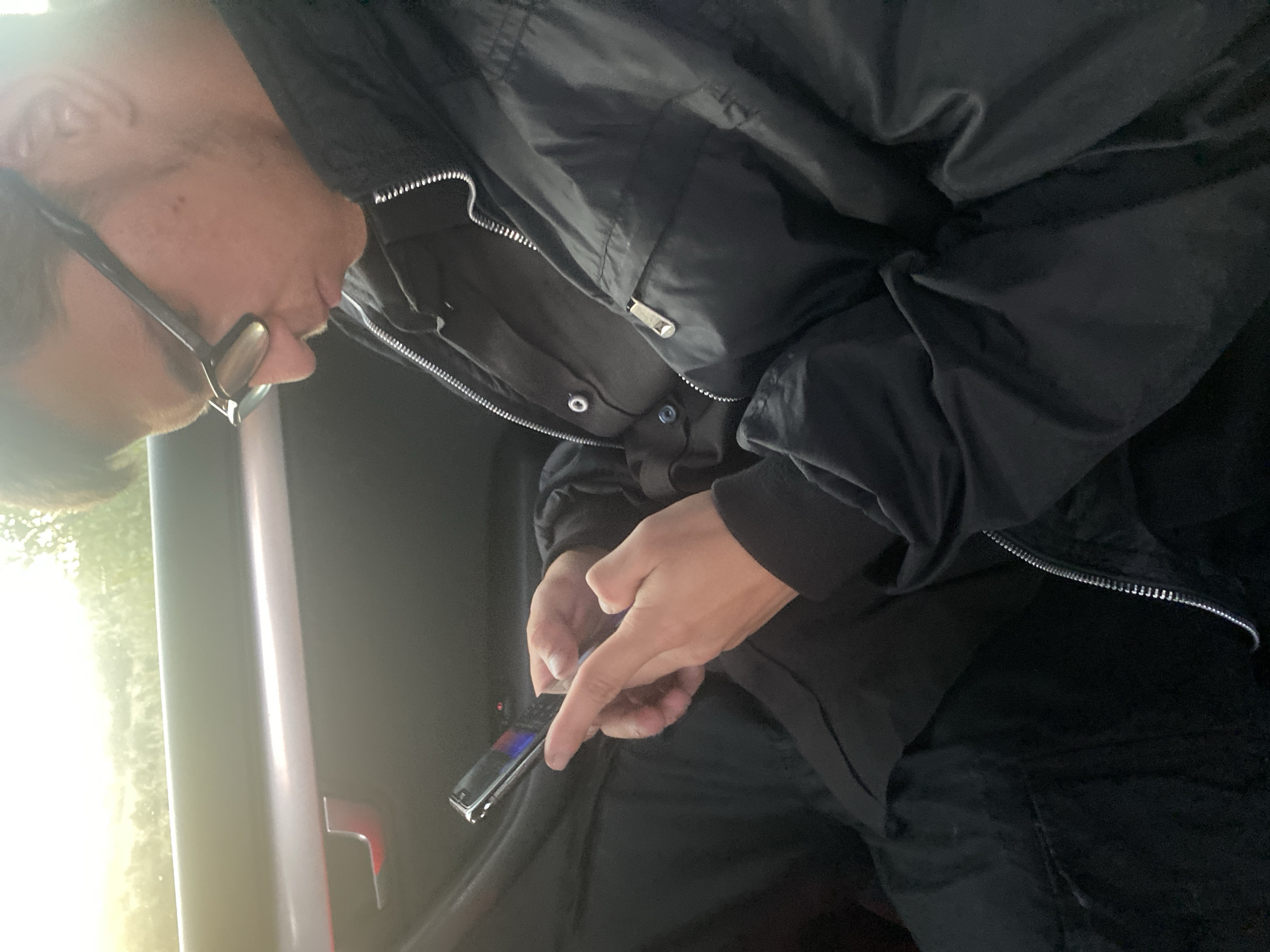 In the city of Ablanitsa, Bulgaria, a person wearing glasses and a black jacket is seated inside a vehicle, focused on their mobile phone. The individual is holding the phone in their hands, and their fingers are visible on the screen, suggesting they are actively using the device. The background shows the interior of the car, including the door handle and a glimpse of the outdoors through the window. The lighting suggests it might be daytime, possibly late afternoon due to the warm, bright light filtering in. The overall scene depicts a moment of personal activity within a vehicle.
