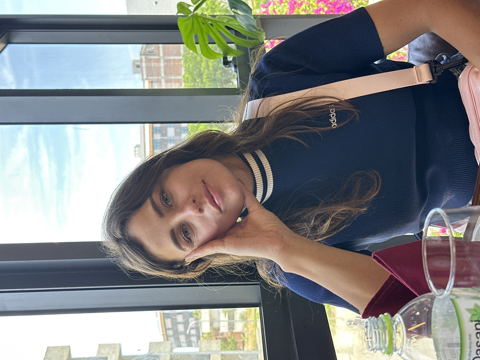 A woman, with long brown hair and green eyes, rests her head on her hand, looking directly at the camera with a gentle smile. She is wearing a dark blue sweater with white stripes around the neckline and sleeves. A light pink strap is visible across her chest. She is seated near a window, with a lush green plant and pink flowers visible outside. The scene appears to be indoors, possibly a cafe or restaurant, given the presence of a glass and a partially visible bottle of Dasani water on a table. The time of day is likely daytime, with natural light streaming in through the window. The woman's pose and expression suggest a relaxed and contemplative mood. Visible text on the bottle reads "Dasani". The location context is Đà Nẵng, Vietnam.