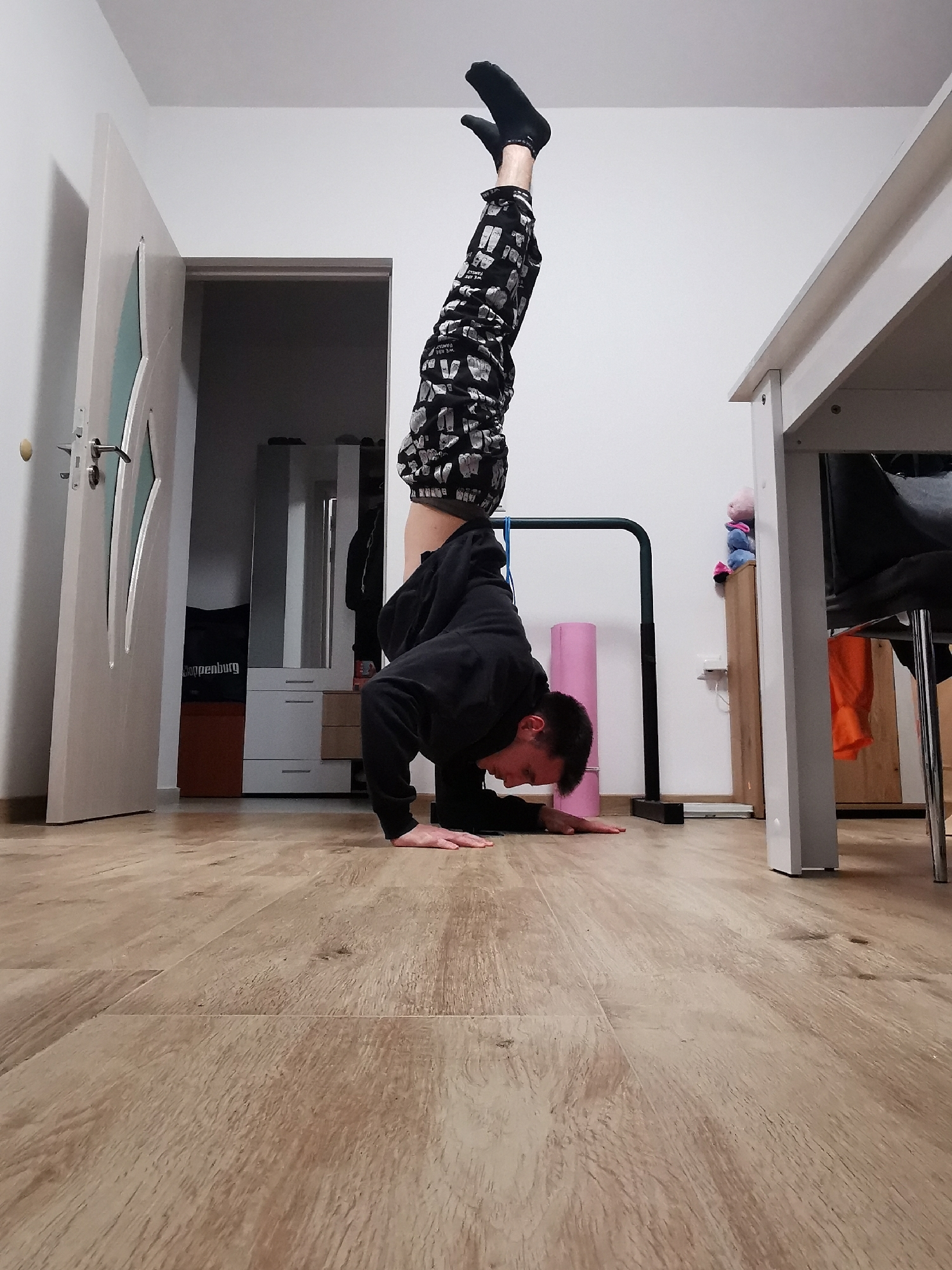 A person is performing a handstand in a room with light wooden flooring. The individual is wearing black socks and patterned black pants with white designs. They are also wearing a black long-sleeved shirt or sweatshirt. The person's head is down towards the floor, with their hands flat on the floor to support their weight.
Behind the person, there is a partially open white door, and further back, a dresser with a mirror. To the right of the person, a white table with a dark chair is partially visible. Also, behind the person, there is some exercise equipment.
Based on the interior, this is likely a residential space, and there is no information indicating it is specifically located in Burgas, Bulgaria.