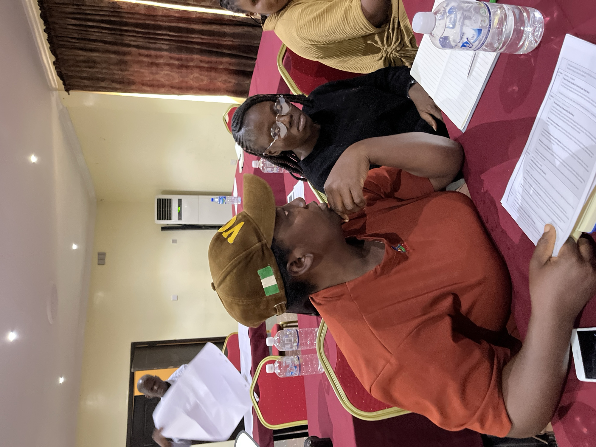 The image depicts three individuals seated at a red-clothed table in an indoor setting, possibly in Gura Top, Nigeria. One person is wearing a brown cap with a Nigerian flag emblem and an orange shirt. Another person is wearing glasses and a black shirt. The third person, partially visible, is wearing a yellow top. Documents, water bottles, and notebooks are on the table. The background shows a wall-mounted air conditioner and a partially visible doorway with a person holding a white sheet of paper. The walls are cream-colored, and brown curtains are visible in the background.