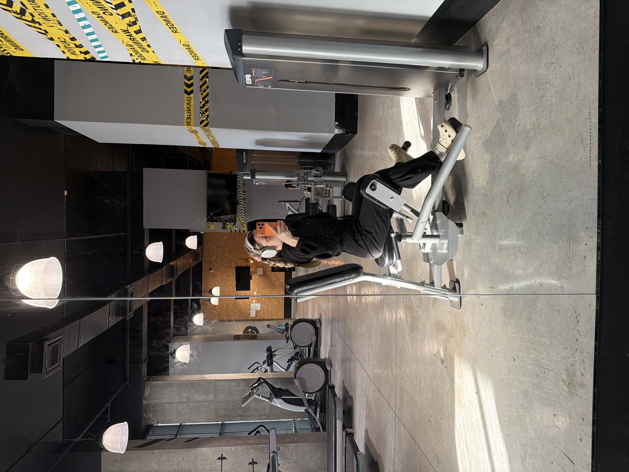 The image shows a woman using a leg press machine at a gym. She is wearing black clothes and beige Crocs, holding a phone in her hand, and listening to headphones. The gym appears modern, with gray concrete floors, black ceiling elements, and rows of cardio machines visible in the reflection of a mirror. The ceiling features white dome-shaped lights and yellow warning tape. The city of Tel Aviv, Israel is not evident in the image.
