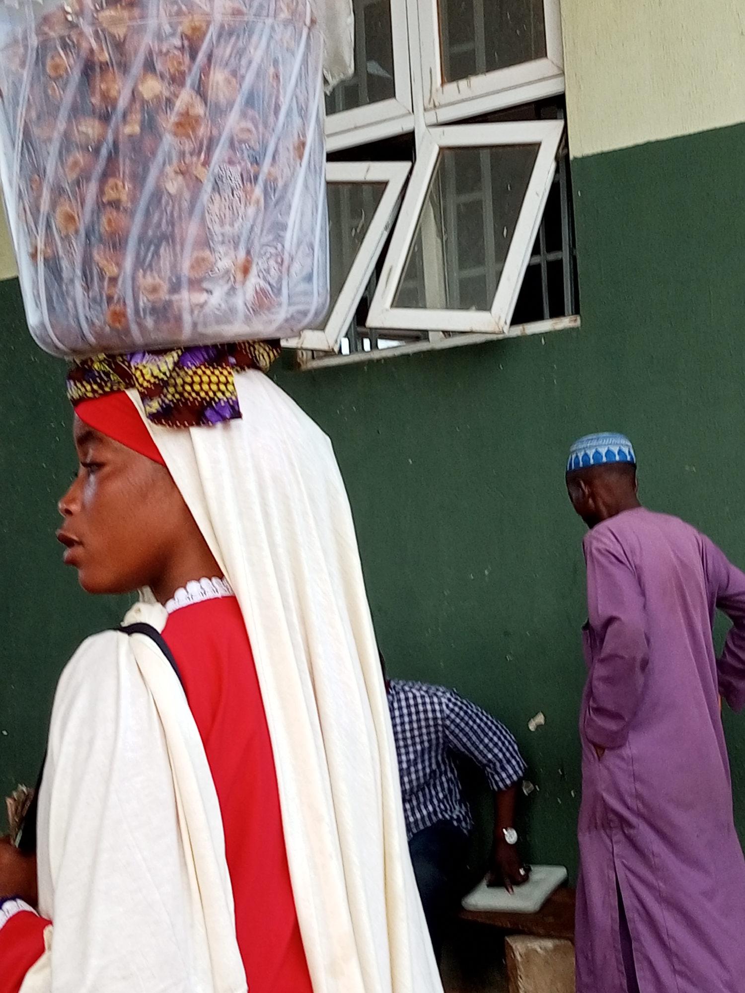 In this image, a young woman is seen in profile, carrying a large, clear plastic container filled with dried dates on her head. She wears a red garment with a white covering draped over her shoulders and head, adorned with a colorful patterned headwrap. To her right, a man in a purple robe and a blue patterned kufi cap stands facing away from the camera. Behind the woman, a person wearing a blue and white checkered shirt is visible, leaning over a small white object on a wooden surface. The scene appears to be outdoors, with a green wall and a window with white frames in the background. The lighting suggests it could be daytime. The overall atmosphere seems to be that of a marketplace or a street scene. There is no visible text in the image.