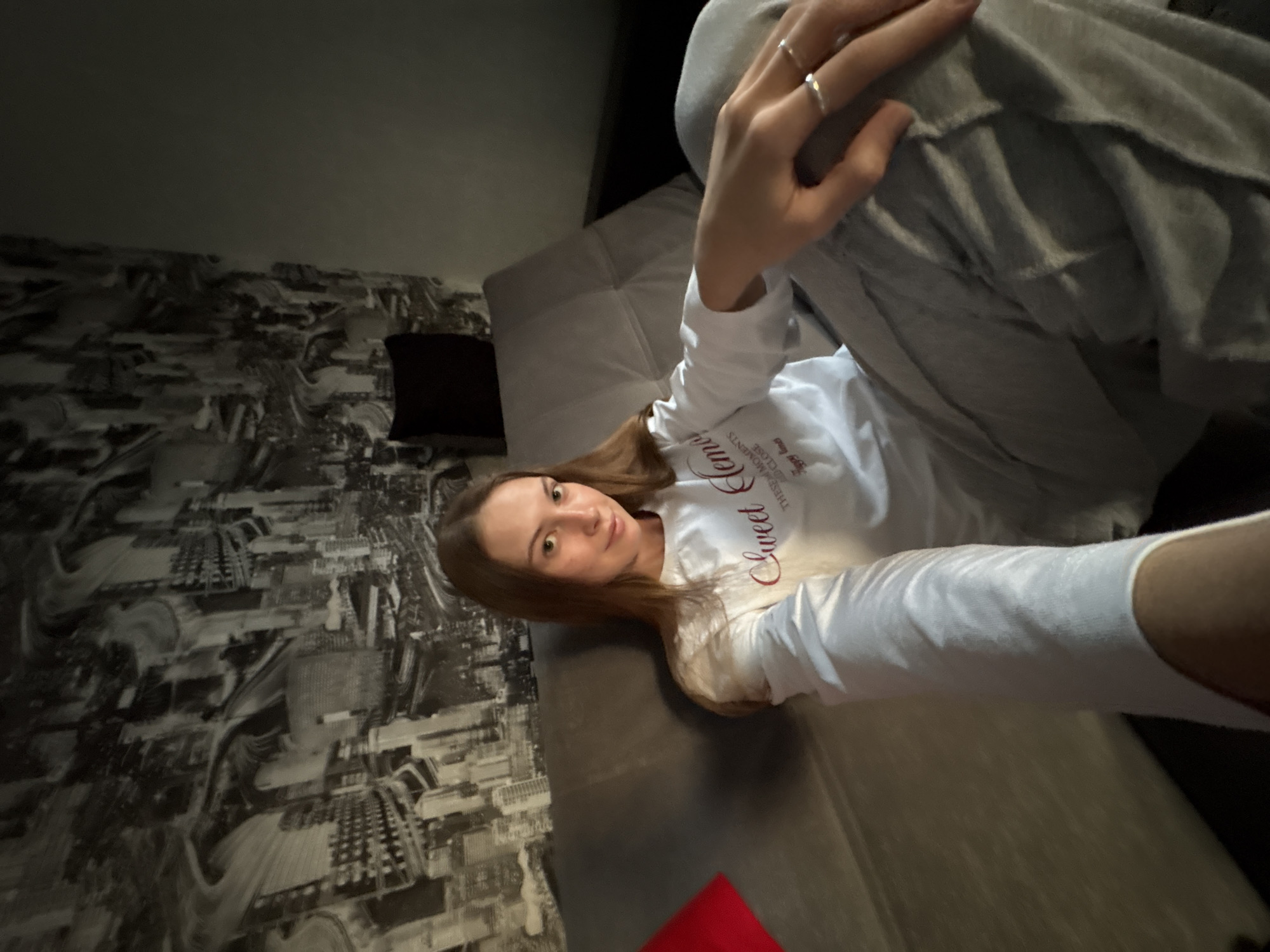 A young woman with long brown hair is sitting on a sofa in a dimly lit room. She is wearing a white long-sleeved shirt with text that appears to say "Sweet Dreams" and gray sweatpants. The background features a black and white mural of a city skyline. A red pillow is visible to the left of the woman. The overall scene suggests a casual and relaxed atmosphere. The location is not identifiable as Novaya Ivanovka, Russia based on the visual information provided.