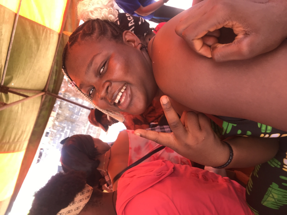 This image depicts a group of individuals in Akpaka Forest Reser, Nigeria, beneath an orange and olive green fabric canopy structure. In the foreground, a young adult female with dark skin and braided hair, interspersed with lighter strands, is shown smiling directly at the camera, revealing teeth. She wears small dark hoop earrings. Her left hand displays a V-sign, while her right hand forms an 'OK' gesture, resting on her right bicep. A dark bracelet is visible on her left wrist. Behind her and to the left, another individual is seen from the back, wearing a red top and a patterned headband. Further back, another person with a dark head covering and a light-colored patterned top is present. Additional individuals are partially visible in the background, including one wearing a red cap and another with light, curly hair. A person in a blue top with white lettering is partially visible on the far right. The background behind the individuals includes a light-colored, textured wall or structure. The scene is illuminated by natural light.