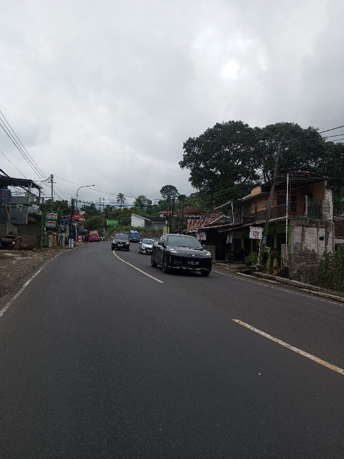 This image depicts a two-lane asphalt road curving to the left, situated in the city of Kodarapalli, India, under a heavily overcast sky. The road features a solid white line at its right edge and a broken yellow center line.
**Vehicles:**
A black SUV, bearing the license plate "B 2471 TMT," is prominent in the mid-foreground, moving towards the viewer on the right side of the road. It is followed by a silver sedan/hatchback, a dark-colored sedan/SUV, and a blue van/minibus, all traveling in the same direction. Further down the road on the left, a pink vehicle is visible.
**Right Side of the Road:**
A two-story building with a light brown upper facade and a light green lower section stands immediately adjacent to the road, featuring a small balcony on the upper level. Signs attached to this building include text that appears to read "WARUNG" and "AYAM GORENG." A large, dense green tree dominates the background behind this structure.
**Left Side of the Road:**
Multiple small structures, some with corrugated metal roofs, line the left side of the road. Utility poles with a dense network of overhead wires are visible. A blue container or water tank rests on a platform. Signs, including one displaying the word "Curuy," are affixed to these structures.
**Environment:**
The background features rolling hills covered in lush green vegetation and trees, including some palm trees. The sky is completely covered by thick, grey clouds, indicating an overcast condition with diffused light. No individuals are distinctly visible in the scene.