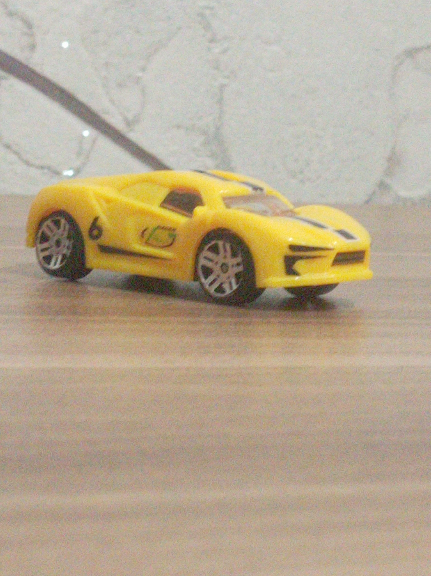 The media file shows a close-up, low-angle shot of a yellow toy sports car. The car is the sole object in the foreground, positioned on a light brown, textured surface that appears to be a table or floor. The background is a lighter, mottled gray surface with a diagonal dark gray stripe and some bright, reflective spots, possibly suggesting a wall with some decorative elements or lighting. The car has black wheels with silver rims, and prominent racing stripes on its front. The number '6' is visible on the side of the car, along with a stylized green logo. The lighting suggests an indoor setting, with no clear indicators of time of day or weather. The image is slightly blurry, indicating a focus on the toy car itself. There is no visible text other than the number '6' and the logo on the car. The context of Baku, Azerbaijan, is provided, but no specific visual cues within the image itself confirm this location.
