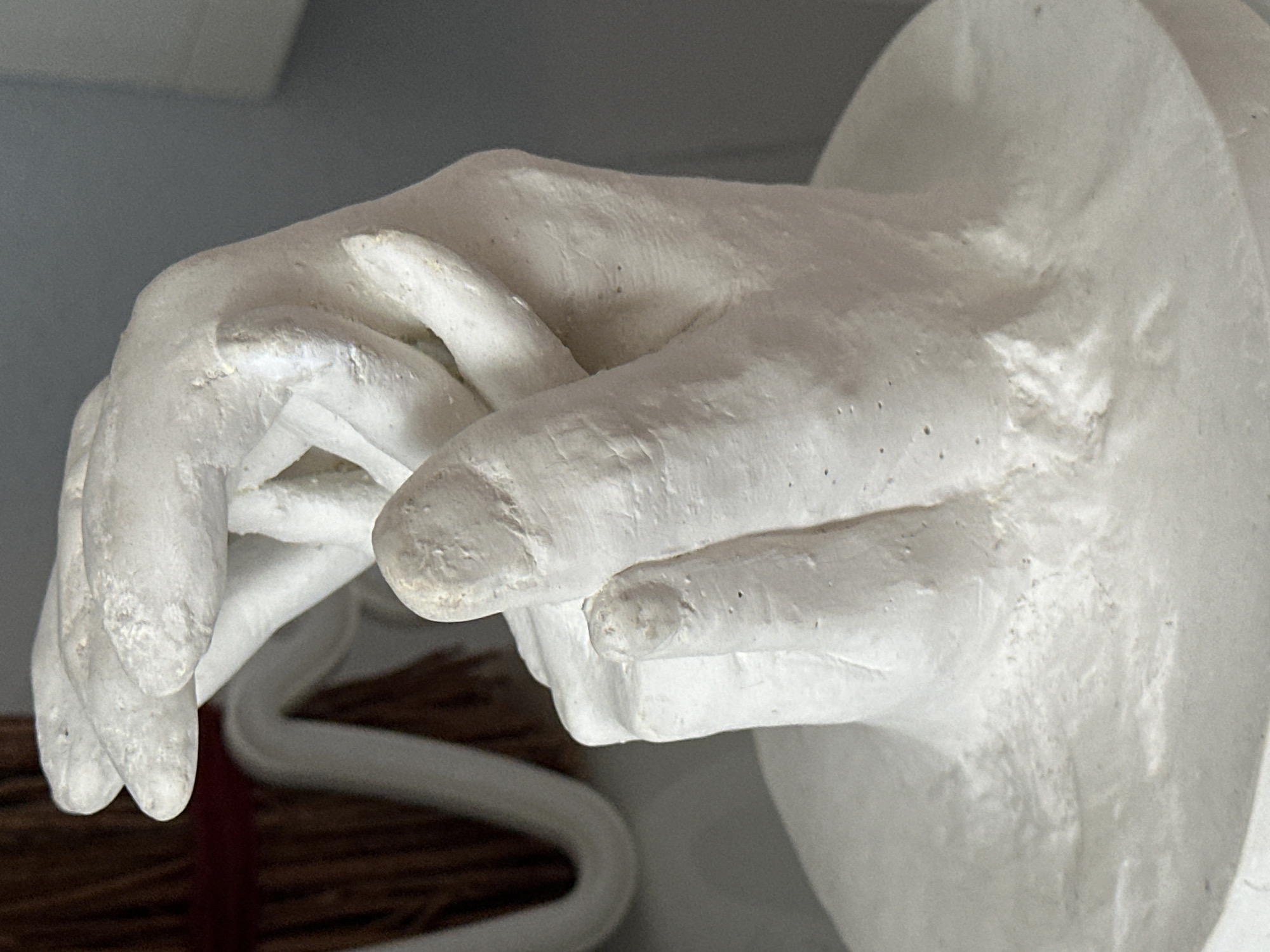 The image displays a close-up, frontal view of a white plaster cast of two hands intertwined. The cast is mounted on a circular white base. The background consists of a dark, textured material on the left and a plain gray surface on the right. The lighting is bright, creating shadows that highlight the textures and contours of the plaster. The object's composition suggests it is a decorative or commemorative piece. The location in Nevinnomyssk, Russia is not discernible from the image itself.