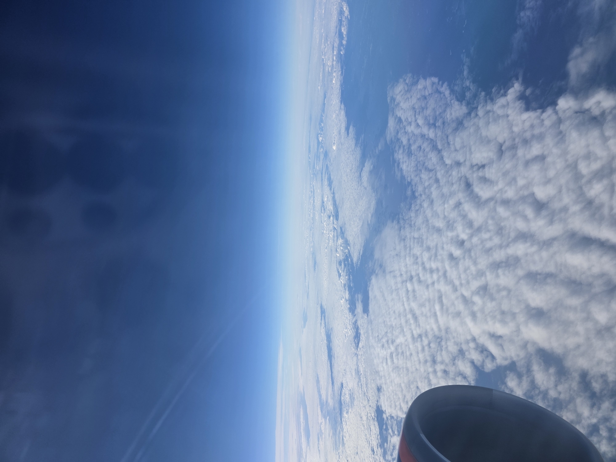 The image is a high-angle view from an airplane window, showing a vast expanse of white clouds below. The clouds are textured and appear to be a thick layer covering the earth. The sky above is a clear, deep blue. The edge of an airplane engine nacelle is visible in the lower left corner of the frame. The image does not contain any information that would allow for the identification of the location in Khimki, Russia.