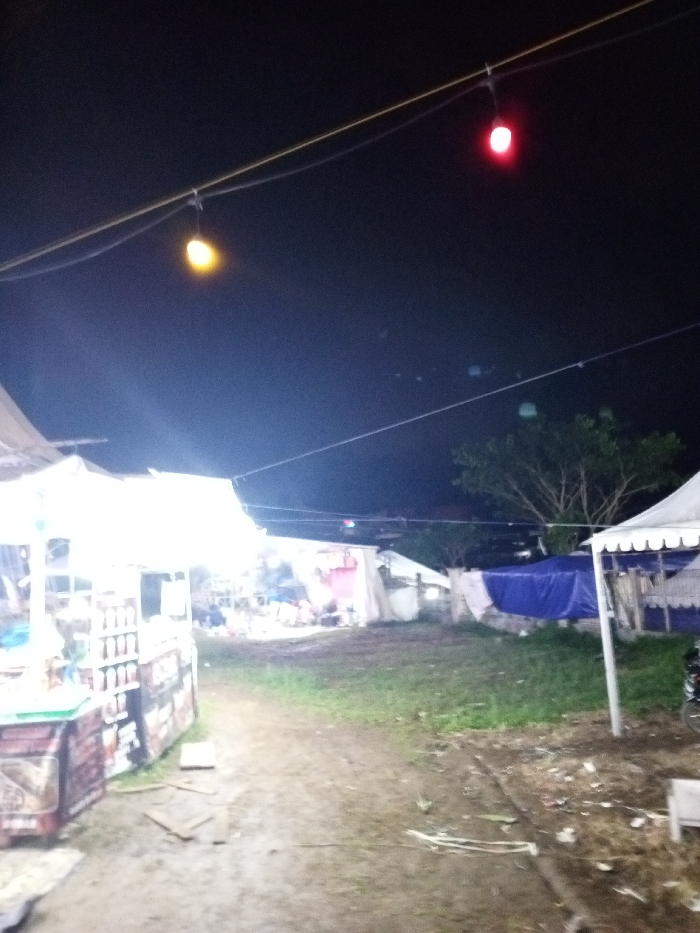 This nighttime photograph captures an outdoor scene, likely a temporary market or gathering, possibly within Khe Sanh Commune, Vietnam. The dark sky dominates the upper portion of the image, punctuated by artificial lights.
In the foreground on the left, a brightly illuminated stall with a white canopy stands out. The intensity of its light source creates an overexposed effect, obscuring precise details within the stall, though rows of bottles or jars can be faintly seen, suggesting it might be a beverage or food vendor. The ground in front of this stall is a mix of dirt and mud, with some debris visible.
A dirt path or open area extends from the foreground into the mid-ground, leading toward several other temporary structures. These include various tents and makeshift shelters, some covered with white tarpaulins and others with blue tarps, indicating an informal or temporary setup. In the background on the right, dark tree foliage is visible against the night sky. On the far right, part of a white tent and a section of a bicycle wheel can be observed.
Above, two string lights are suspended across the dark expanse. One light on the left glows with a warm yellow-orange hue, while another further to the right emits a distinct red light. The overall illumination is sparse, with the prominent stall on the left providing the most intense light. No individuals are visible in the photograph, lending a quiet atmosphere to what appears to be a communal space or event. There is no discernible text present within the image.