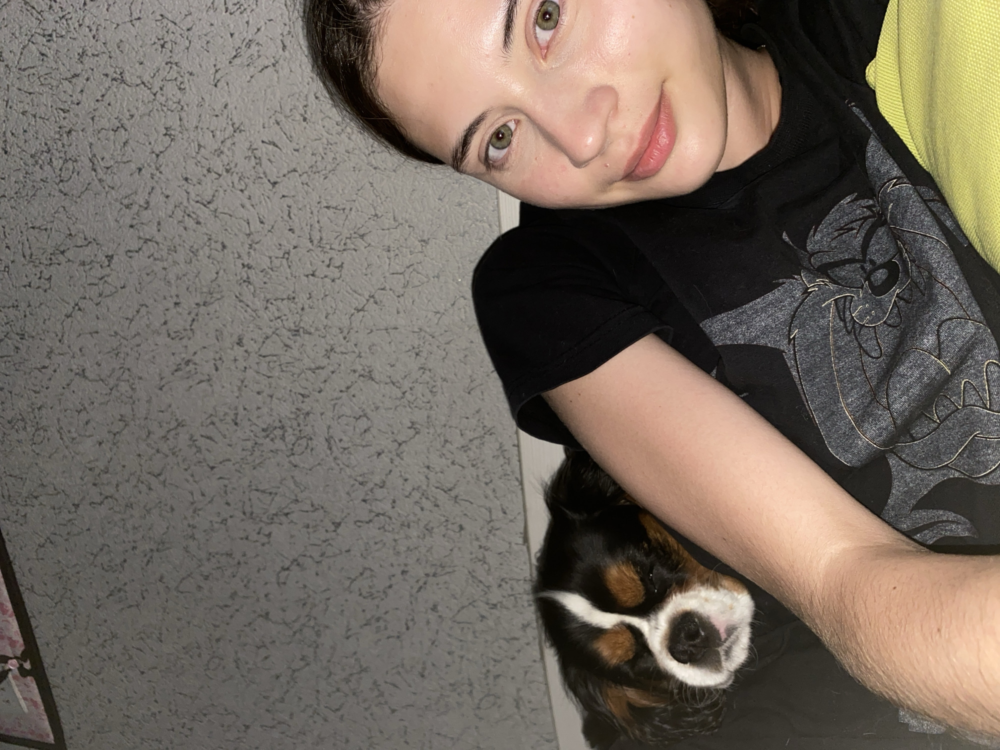 The image is a close-up, slightly angled selfie of a young woman with light skin and green eyes, lying down with her head on a pillow. A small dog, a Cavalier King Charles Spaniel with tri-color markings, is nestled beside her, its head resting on her shoulder. Both the woman and the dog appear relaxed. The background is a textured, grey wall. The woman is wearing a black t-shirt with a graphic print. The scene appears to be indoors, possibly in a home environment. There is no visible indication of the specific location in Votkinsk, Russia, beyond the provided information.