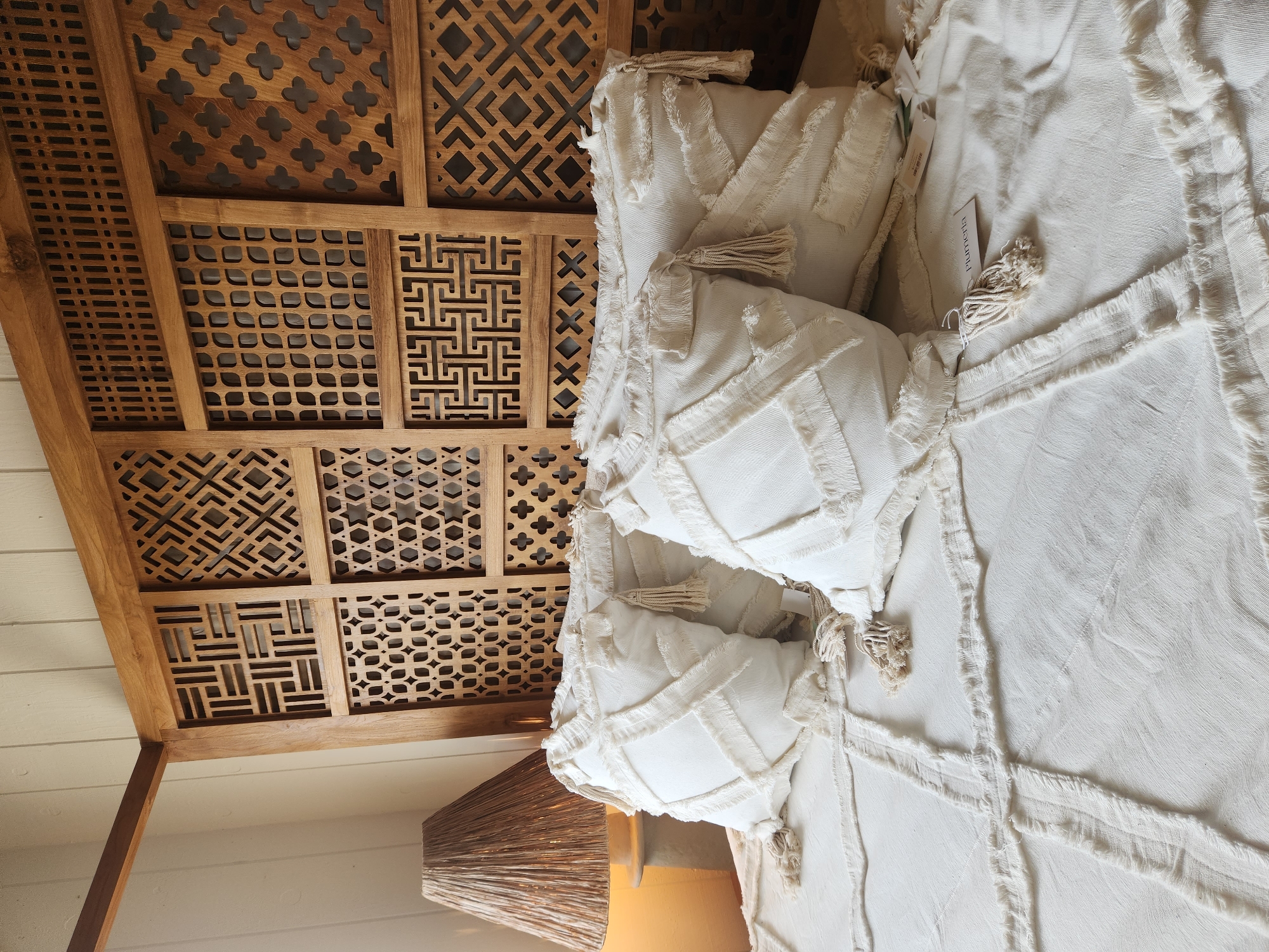 The image shows a close-up view of a bed with white, textured pillows and a white bedspread with fringed detailing. The pillows have a crisscross pattern of fabric strips and tassels. The bed is situated in front of a large, ornate wooden headboard with intricate geometric carvings. To the left of the bed, a lamp with a woven shade is visible. The background appears to be a wall with vertical wooden paneling. The scene is well-lit, with warm lighting, suggesting an indoor setting. The image does not contain any explicit information about the city of Maitland, United States of America, or any specific location within it.