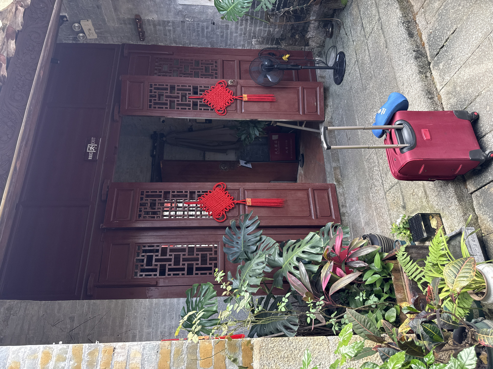 A still life image depicts a doorway adorned with traditional Chinese knot decorations and a suitcase. The entrance features large, dark red wooden doors with intricate lattice work. Red Chinese knots hang on either side of the doorway, one on each door panel. To the left of the doorway, a collection of potted plants with large, green leaves and some with red foliage are arranged on the stone ground. A red suitcase with a blue neck pillow on top sits on the stone pathway in front of the doorway. To the right of the doorway, a black oscillating fan stands on a black base. The wall on the right is constructed from grey bricks, while the wall on the left is a mix of grey brick and rough-textured yellow and grey stone. The ground is paved with grey stone slabs. Above the doorway, an exit sign is visible, indicating the location is likely within a public or commercial space. The scene appears to be an outdoor entrance to a building, possibly in Longgang District, China, given the architectural style and decorations.