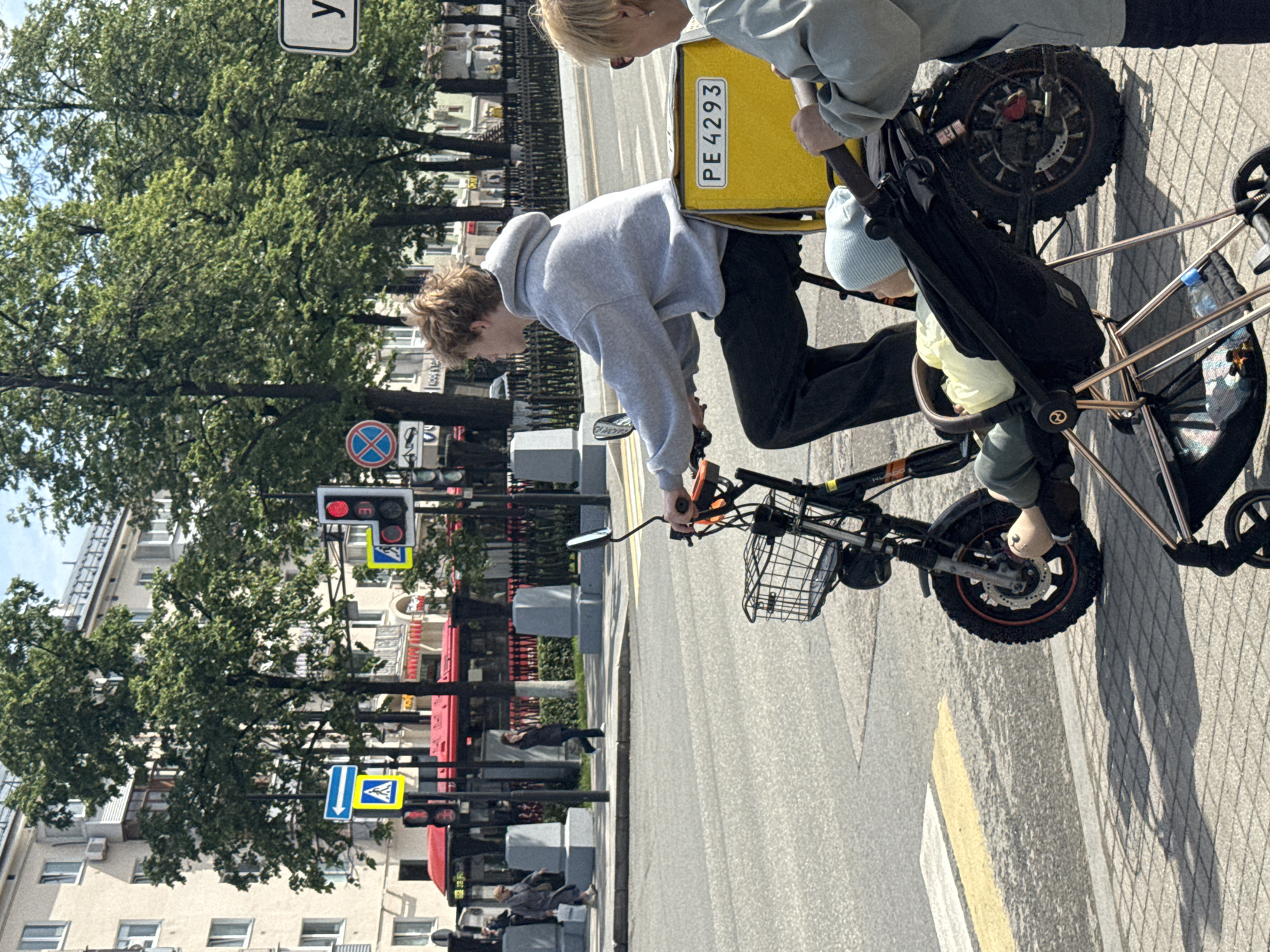 This daytime image, taken in Perm, Russia, depicts a street scene centered on two individuals and an electric bicycle. A person with light, wavy hair, dressed in a grey hoodie and dark pants, is positioned on or next to a black electric bicycle. The bicycle is equipped with a front wire basket and a large, bright yellow delivery box on its rear, which displays the text "PE 4293".
Adjacent to the bicycle, a child, wearing a light blue beanie and a light-colored top, is seated in a dark-colored stroller or child carrier. This carrier appears to be either integrated with the bicycle setup or being pushed alongside it.
The setting is a bustling urban environment with a wide paved road and sidewalk. Numerous mature green trees line the street, providing a natural backdrop against several multi-story buildings typical of city architecture. Visible street furniture includes traffic lights, one displaying a red signal, and various road signs: a partially visible white rectangular sign with the letter "y," a blue circular "no stopping" or "no waiting" sign, and blue and yellow square signs indicating a pedestrian crossing. The bright sun casts clear shadows, suggesting a sunny day. Other pedestrians can be seen further down the street.