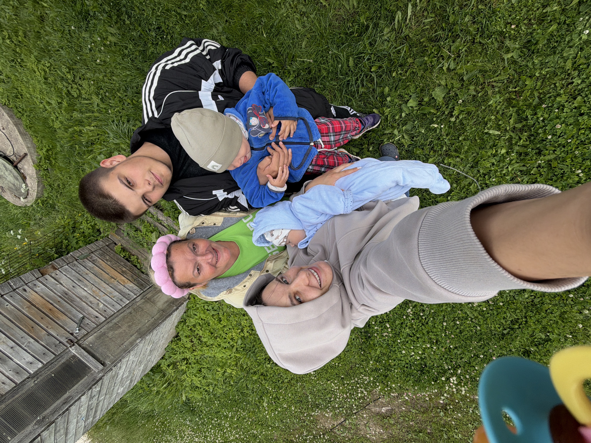 In the city of Andreyevo, Russia, a group of four individuals is captured in a high-angle selfie taken outdoors. The setting is a grassy area, possibly a yard or park, with a wooden structure visible in the upper left corner, resembling a shed or outbuilding.
The individuals appear to be two women, a teenage boy, and two young children. One woman, wearing a light beige hooded sweatshirt, holds a baby wrapped in a light blue swaddle. She is smiling directly at the camera. Another woman, positioned behind her, wears a green shirt and a pink padded headband. She is also smiling. The teenage boy, dressed in a black and white Adidas jacket, is holding a toddler. The toddler is wearing a blue jacket and red plaid pants, with a light grey beanie on his head. His tongue is sticking out slightly. The scene suggests a family gathering or outing.
